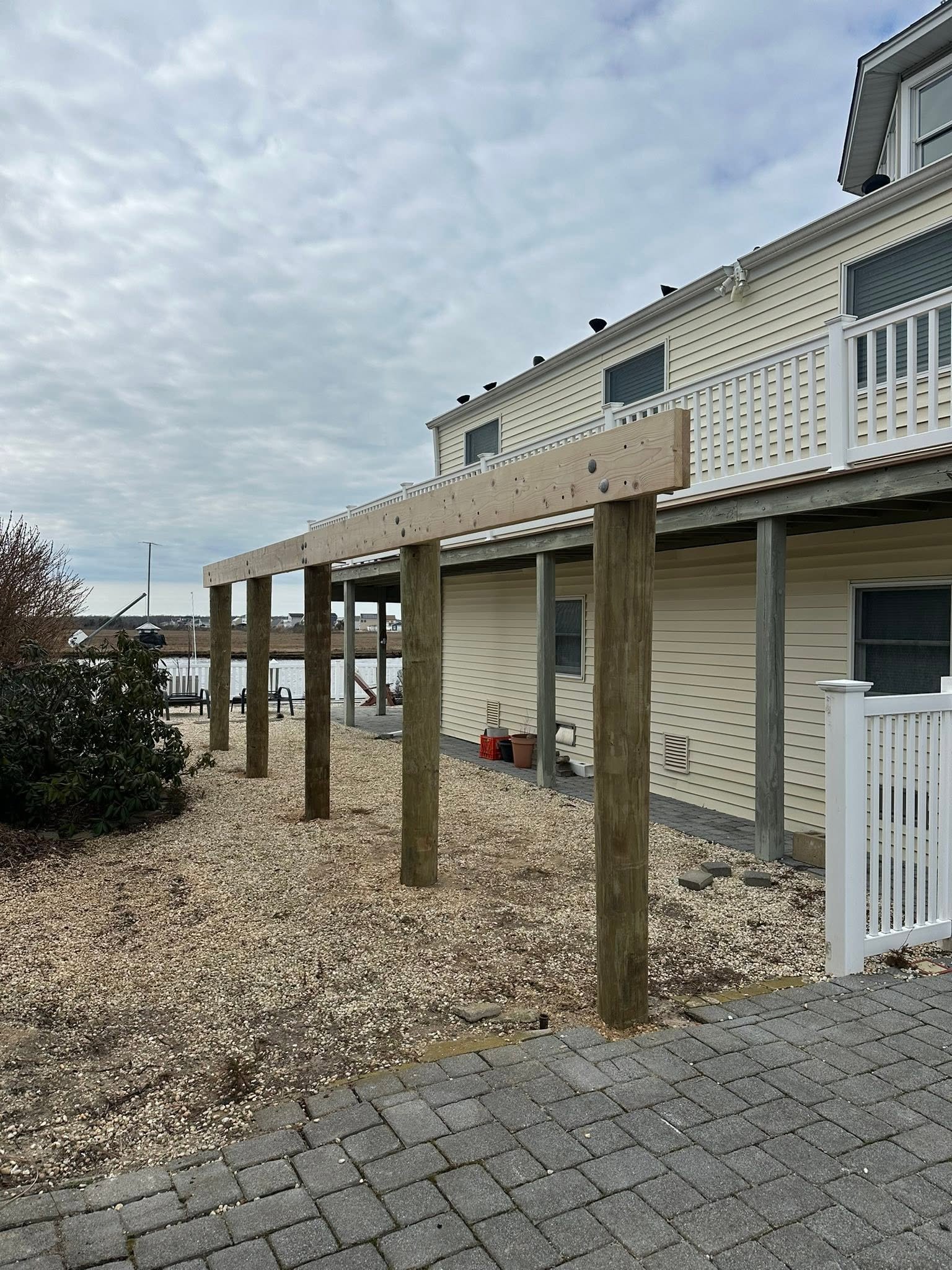 Backyard of a house under construction with wooden posts for a deck and a gravel ground. The house has beige siding, white railing, and multiple windows. Potted plants and outdoor furniture are visible near the house, and the sky is cloudy.