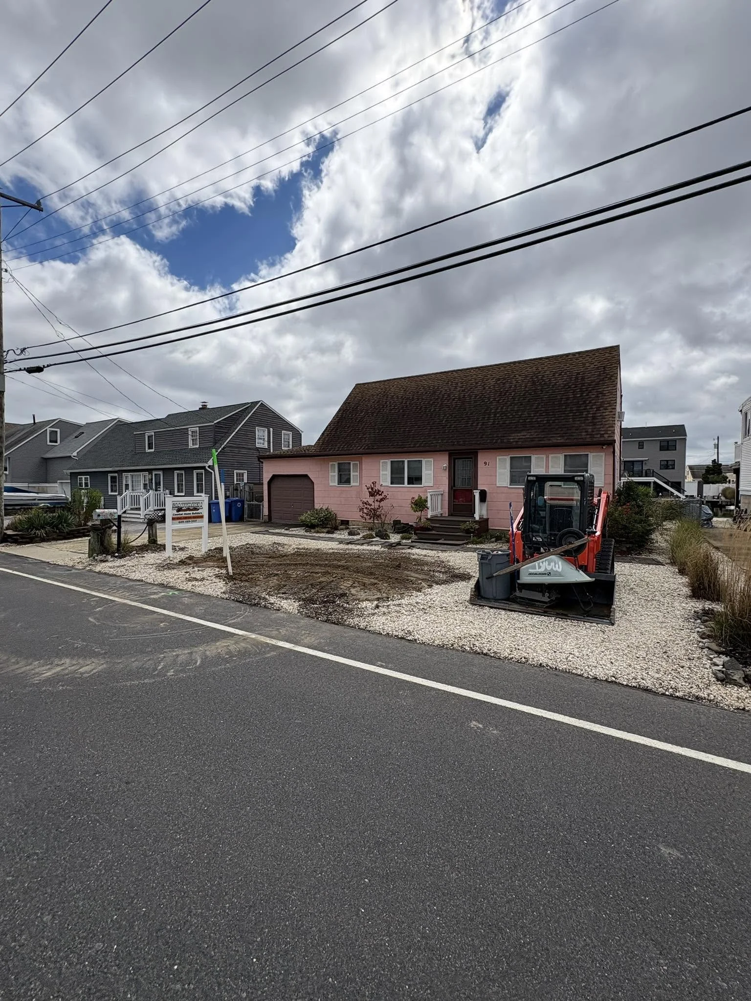 A pink house with a brown roof under a cloudy sky, construction equipment and gravel in the front yard, and neighboring houses visible.