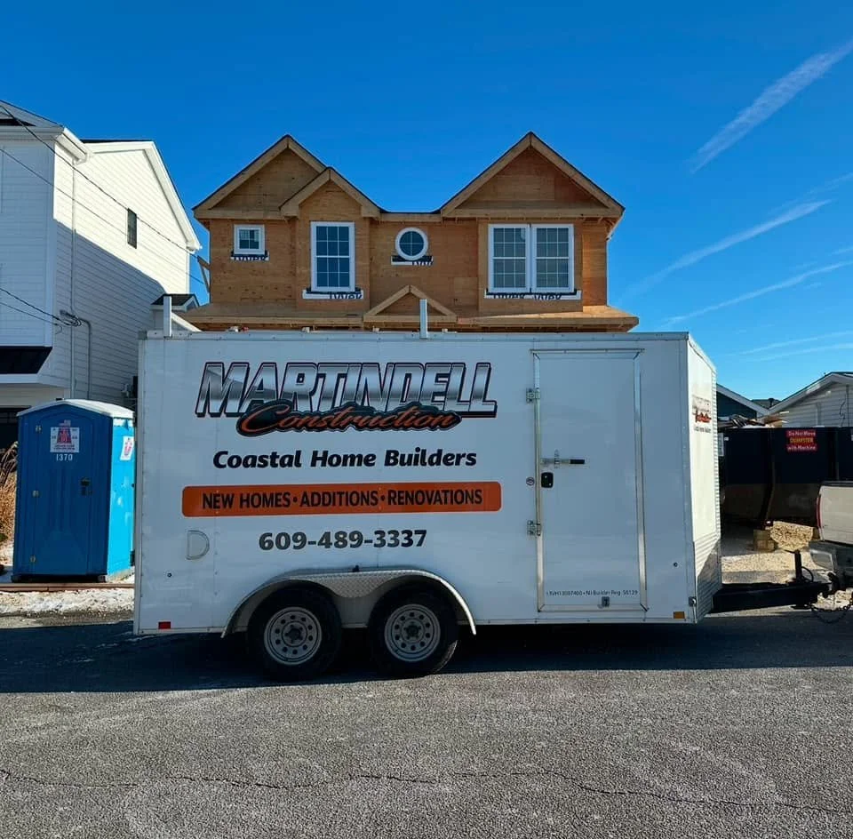 Construction trailer for Martindell Construction, a coastal home builder, parked in front of a house under construction with a partially completed upper story. A portable toilet is nearby under a clear blue sky.