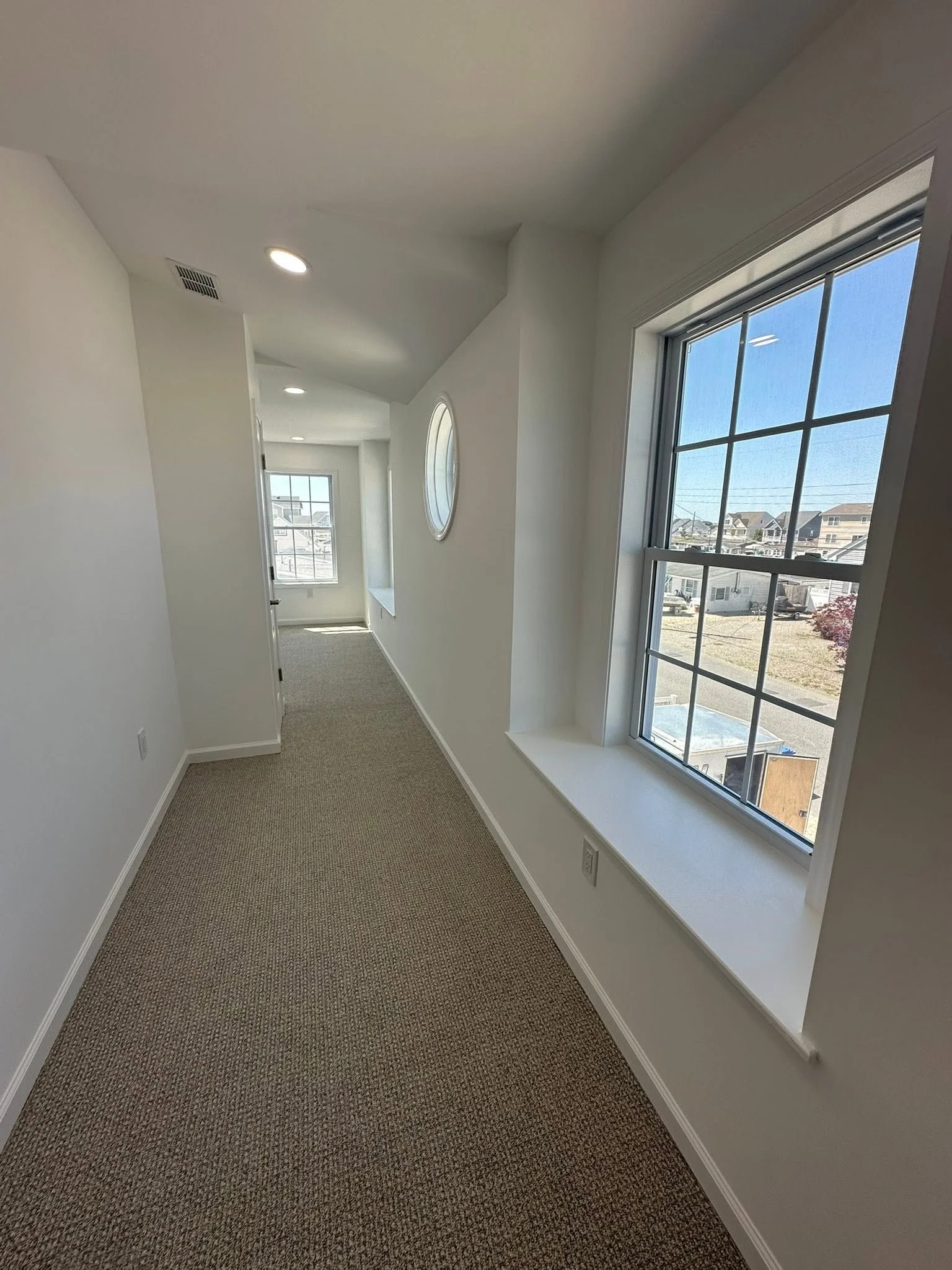 Hallway with large window showing a neighborhood outside, white walls, beige carpet, and a circular window on the wall.