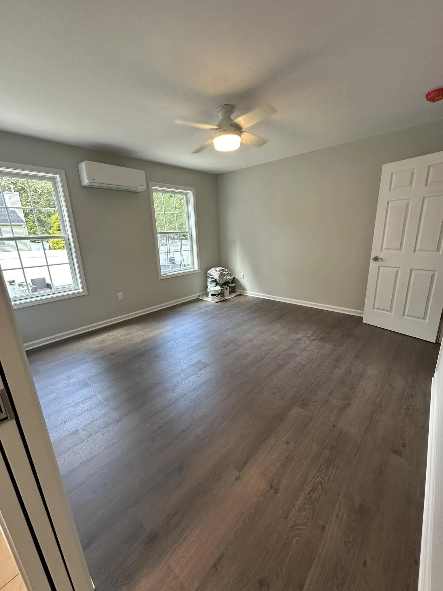 Empty room with two windows, a ceiling fan with light, a white door slightly open, and some paint supplies stacked in a corner.