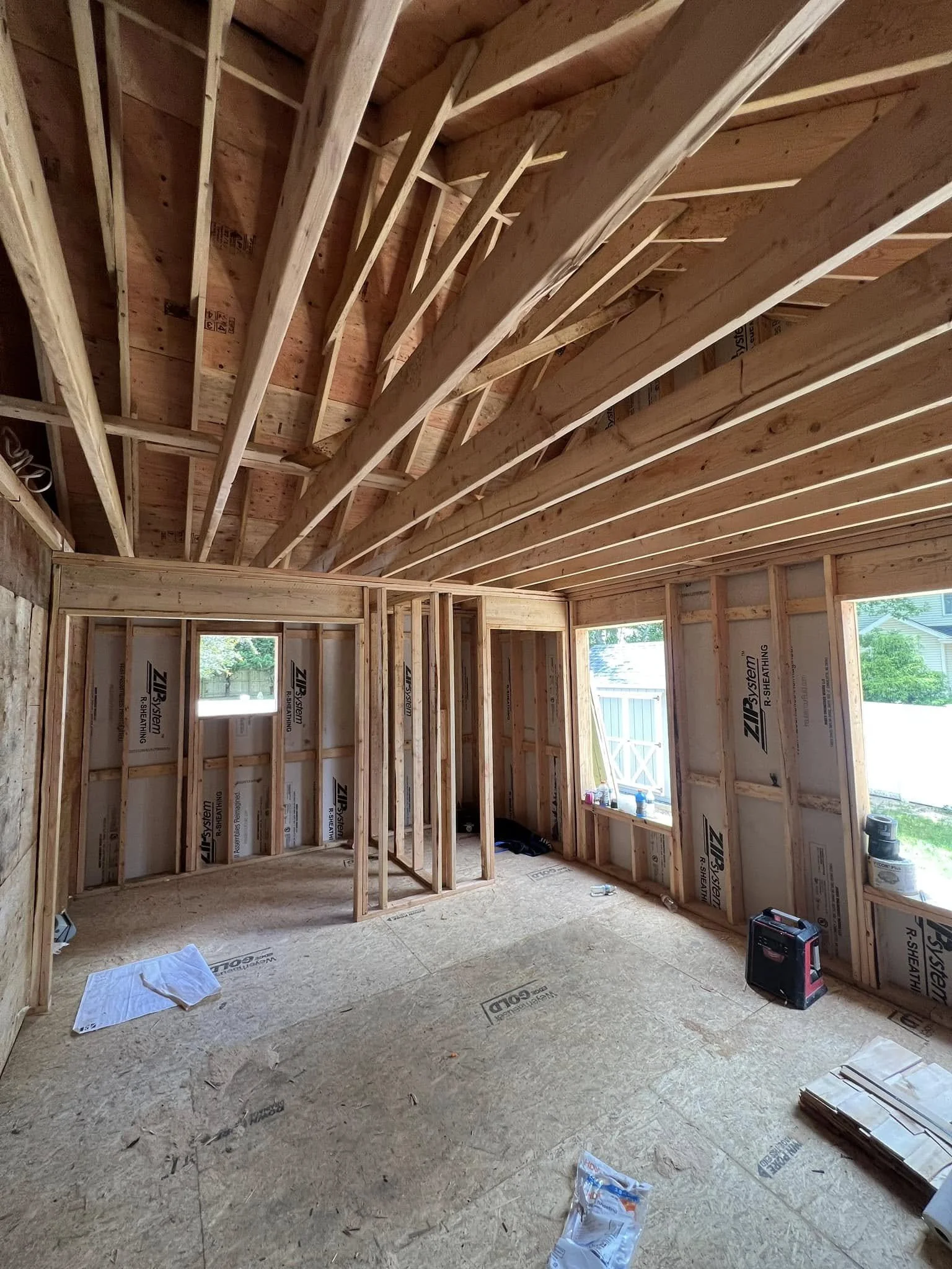 Interior of a house under construction showing exposed framing, some walls with building wrap, and an unfinished ceiling with wooden joists and insulation.