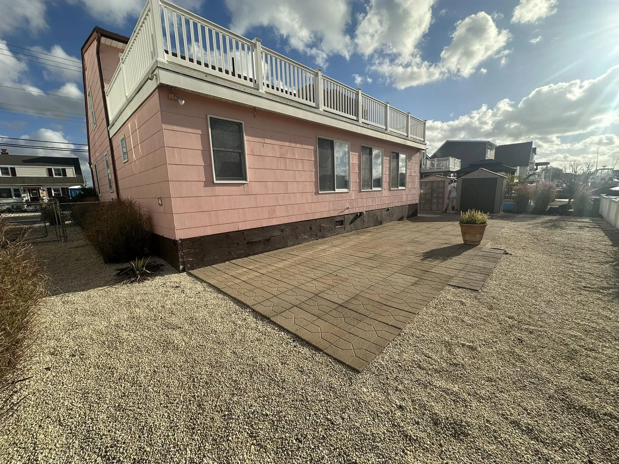 A two-story pink house with a large outdoor deck and white railing, set against a partly cloudy sky and neighboring houses, with a paved patio and gravel yard in the foreground.