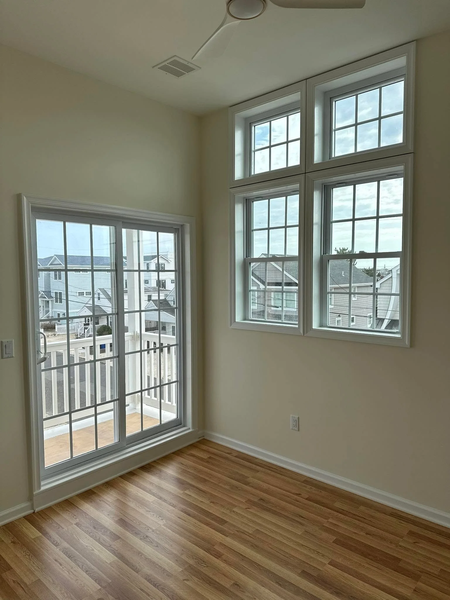 Bright room with large windows and a sliding glass door leading to a small balcony. The room has wooden flooring and light-colored walls.