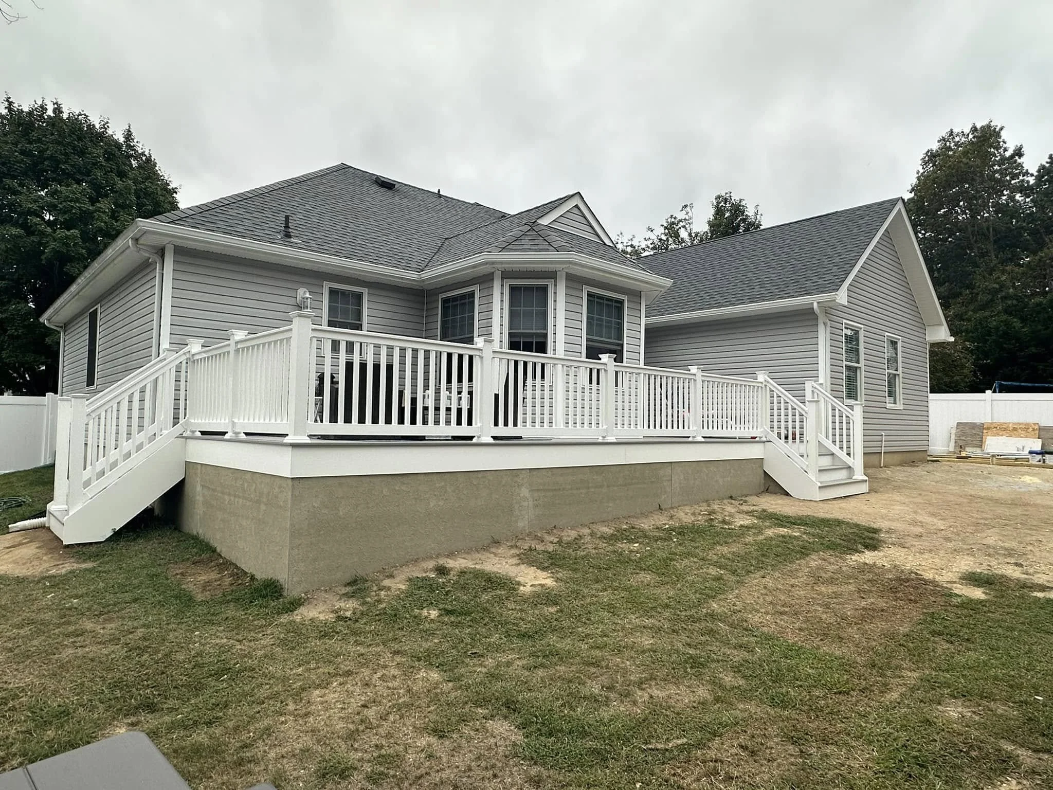 Backyard view of a two-story house with gray siding, white trim, multiple windows, a deck with white railing, and a surrounding white fence, under an overcast sky.