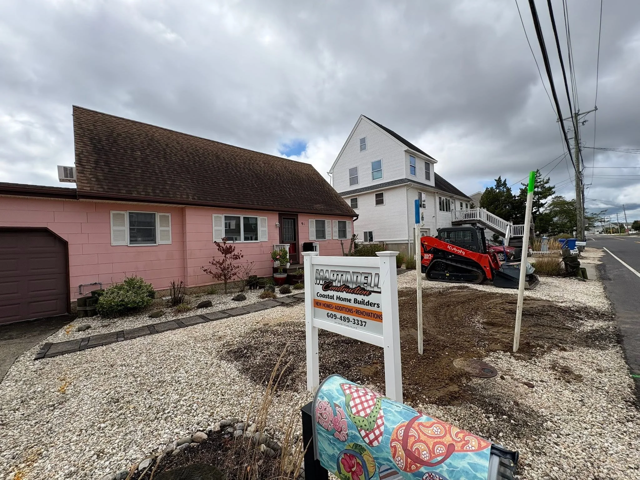 A small pink house with a brown roof next to a larger white house. There's a sign for a construction company in front, a small gravel yard with plants, and a colorful chair in the foreground. Overcast sky with clouds and power lines overhead.