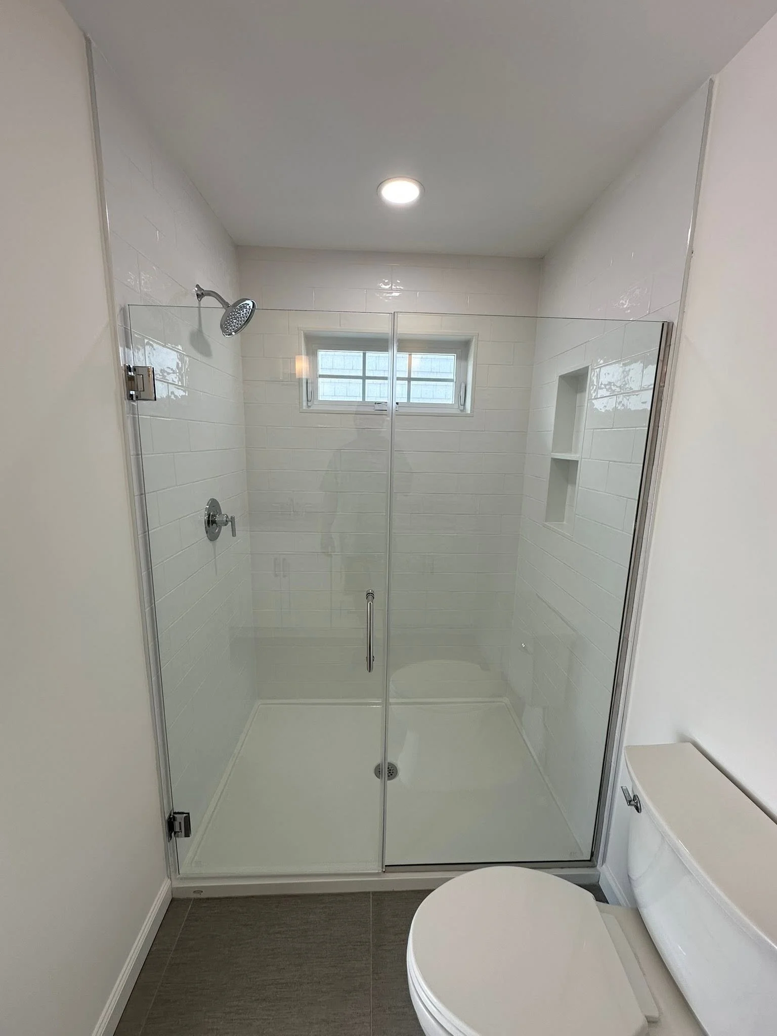Bathroom with a glass shower enclosure, white tiled walls, a small window, and a white toilet.