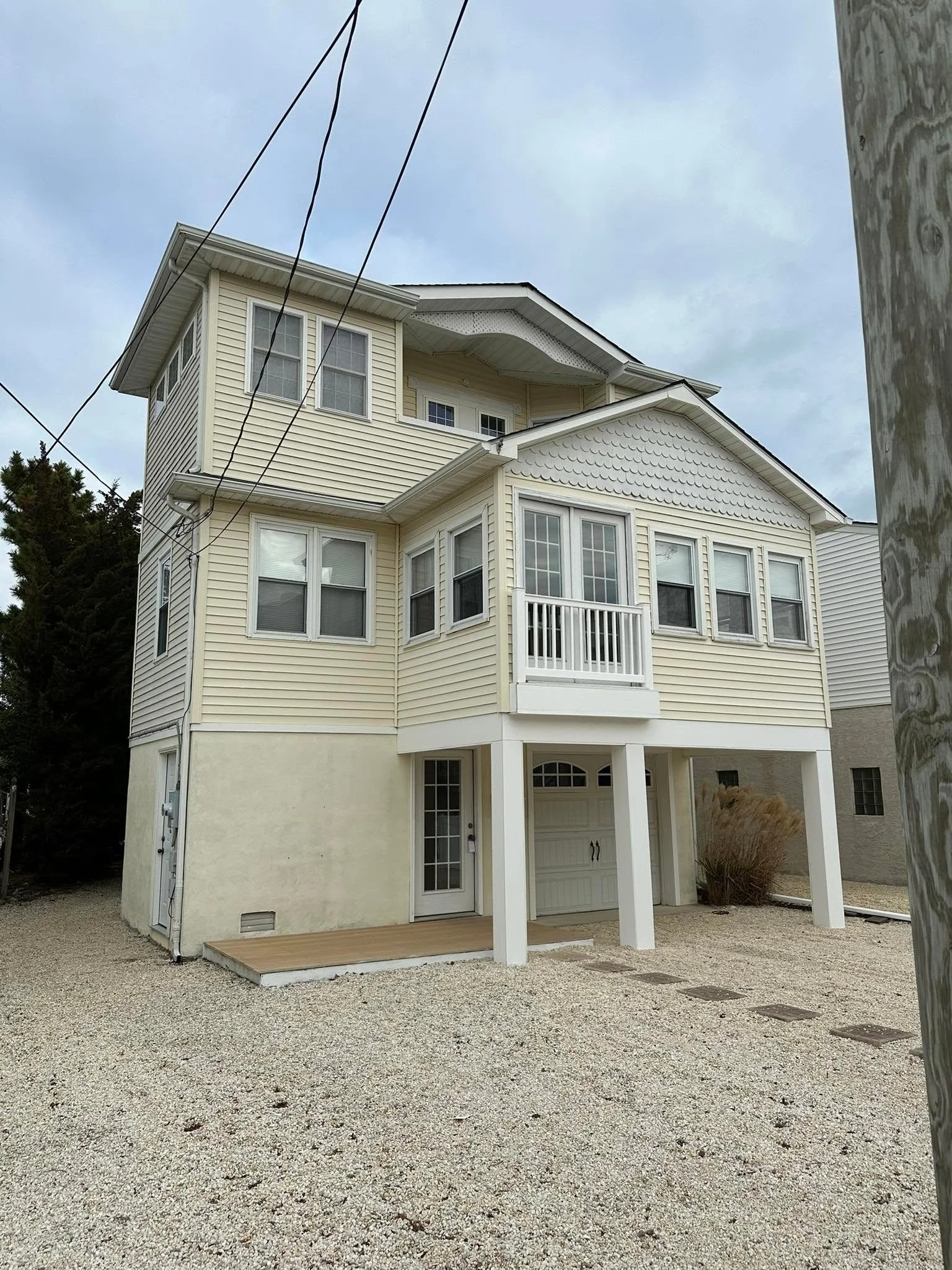 A multi-story yellow house with white trim, a small balcony, and a garage door, standing on a gravel surface with a nearby wooden utility pole and some plants.