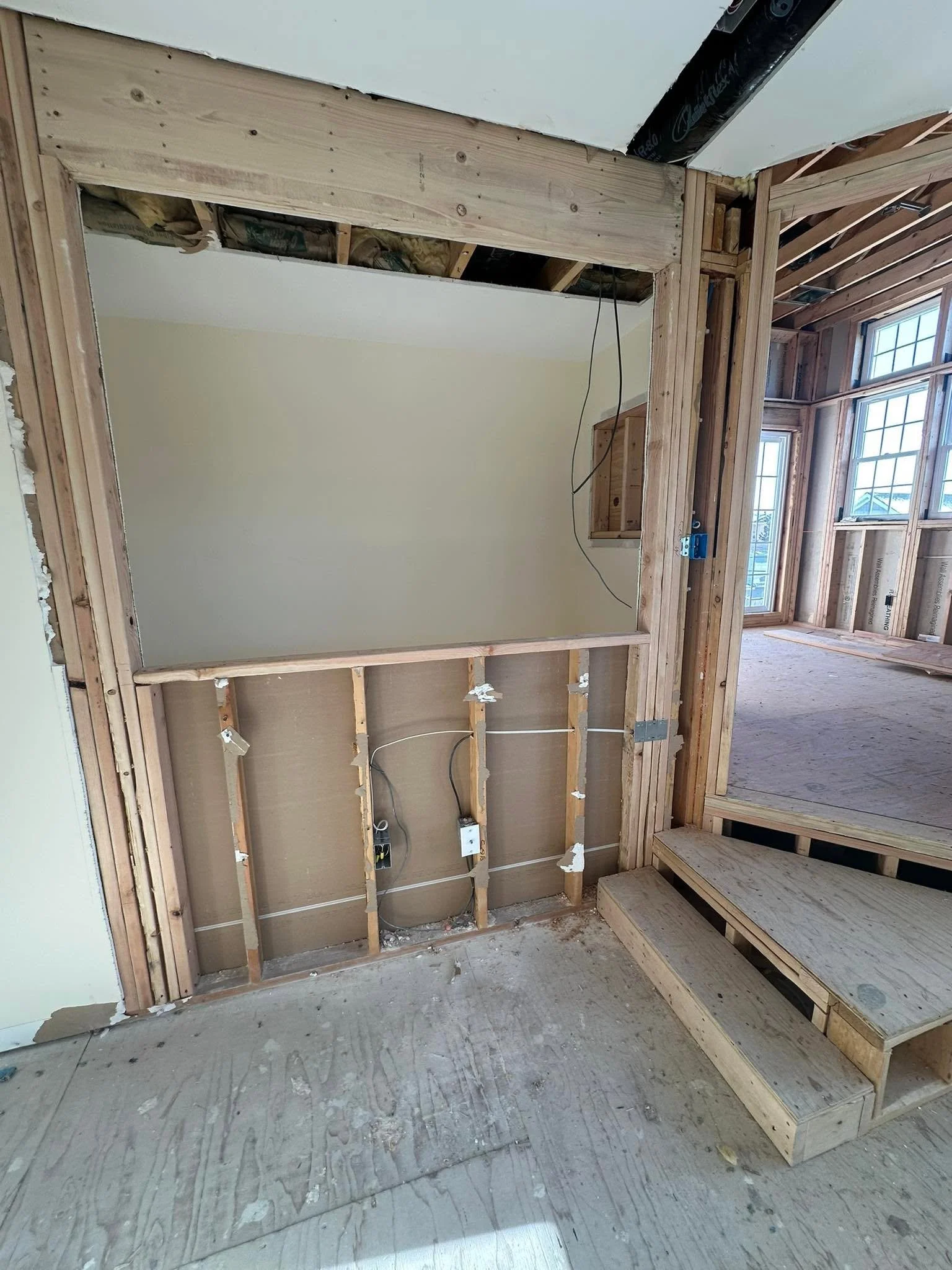 Interior of a house under construction with wooden framing and drywall installed on one wall, two large windows in the background, and a staircase on the right. The floor is unfinished concrete.