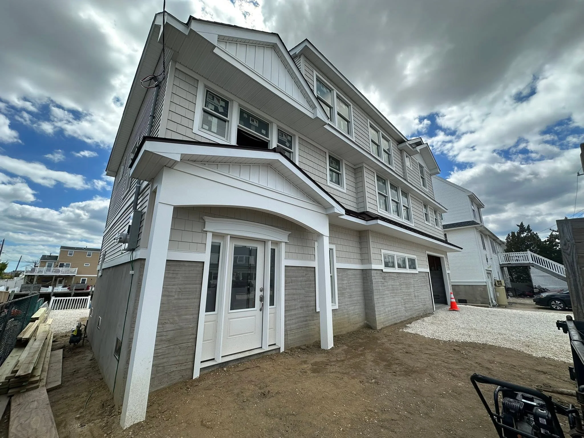A partially constructed multi-story house with white siding, multiple windows, and a small balcony. The area around the house is under construction with dirt, gravel, and building materials visible. The sky is partly cloudy.