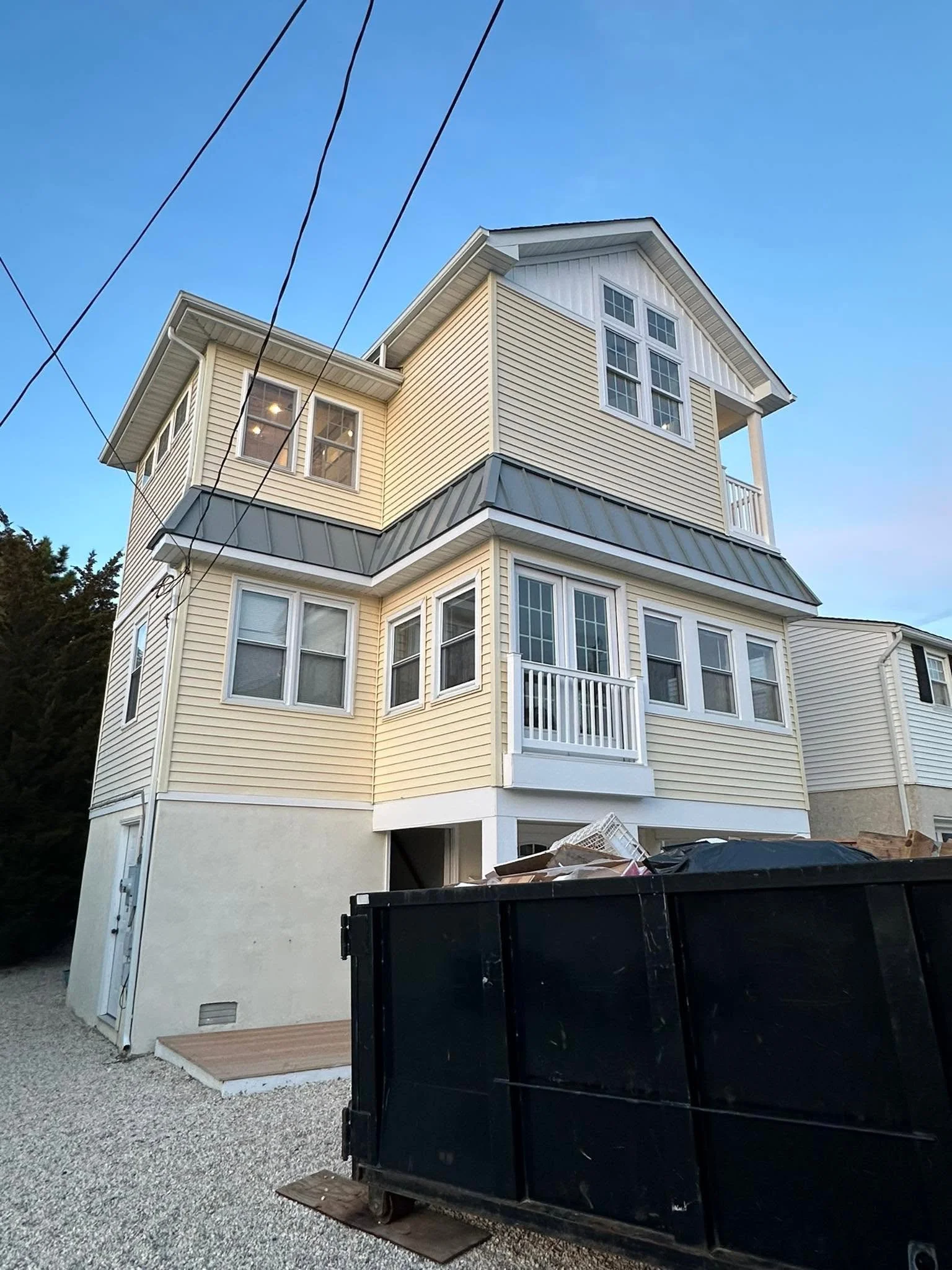 A multi-story house with beige siding and white trim, large windows, a small balcony, and a gray metal roof, under a blue sky. There is a black dumpster filled with debris and an open door area at the bottom of the house.