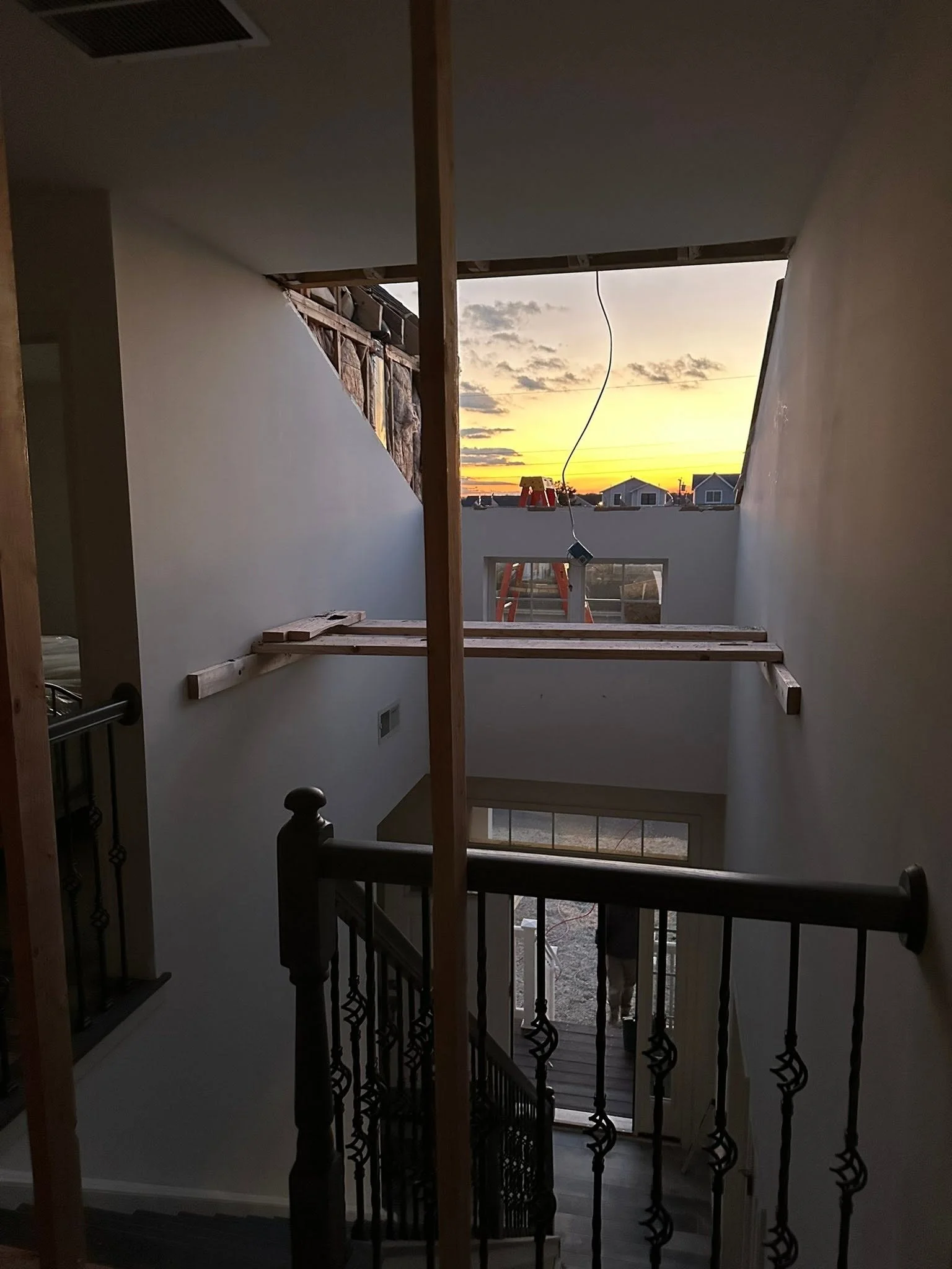 Interior view of a house under construction with a staircase and a large window showing a sunset sky with clouds and houses outside.