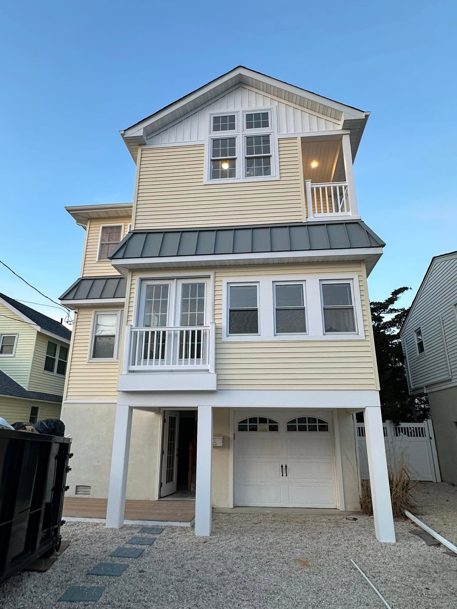 The back of a three-story house with beige and white siding, a garage, and a small balcony on the second floor. There is a gravel driveway and outdoor trash bins on the left side.