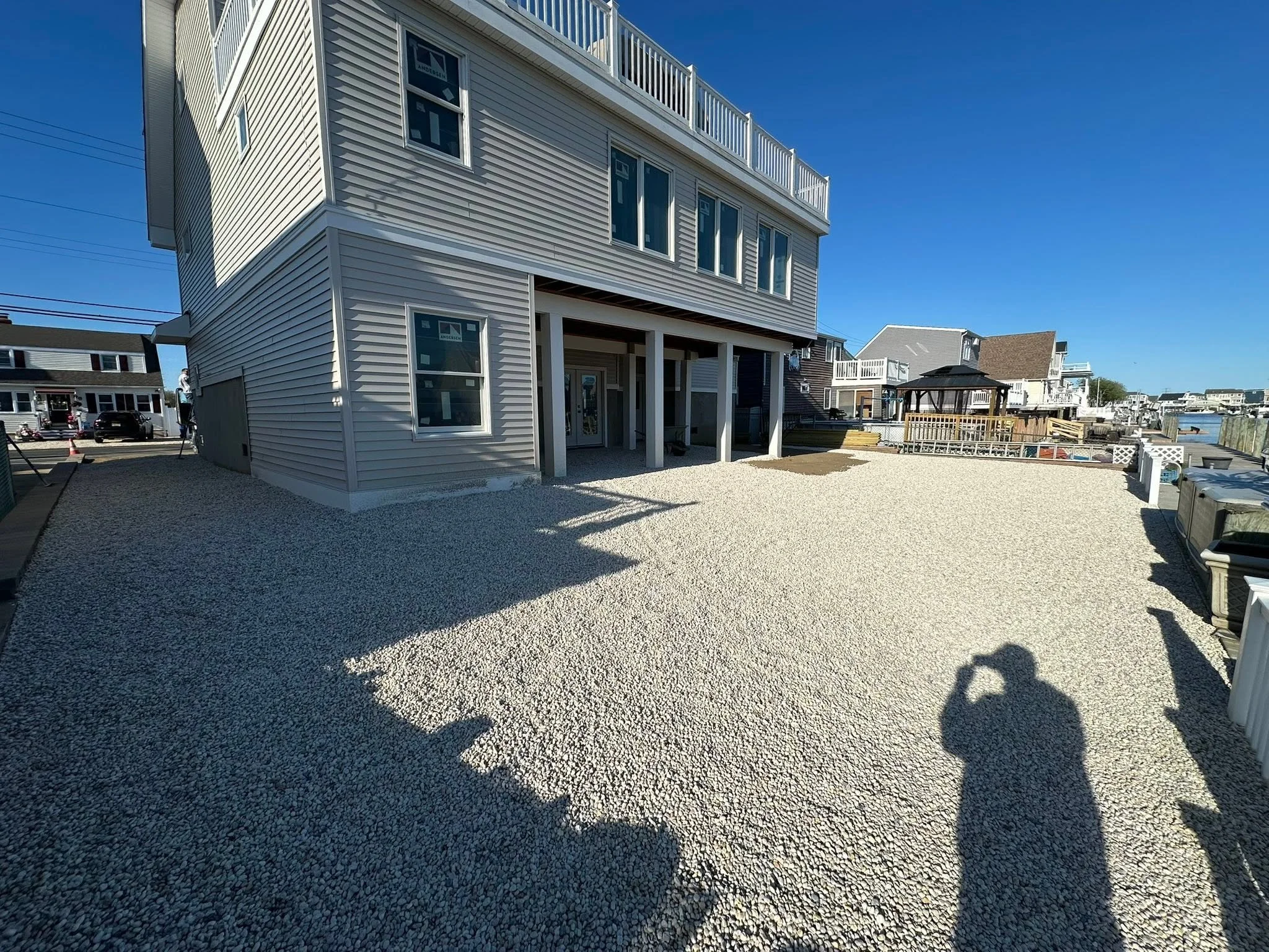 View of a modern multi-story house with vinyl siding, large windows, and a balcony, situated by a waterfront on a clear, sunny day.