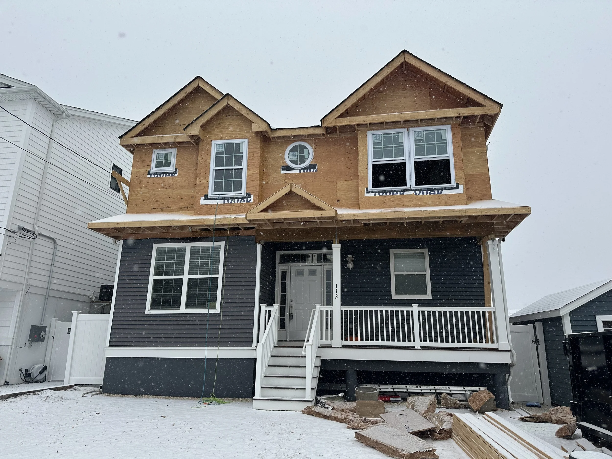 A house under construction with exposed wooden framing, a partial second story, and a front porch. Snow is falling on the yard and surrounding area.