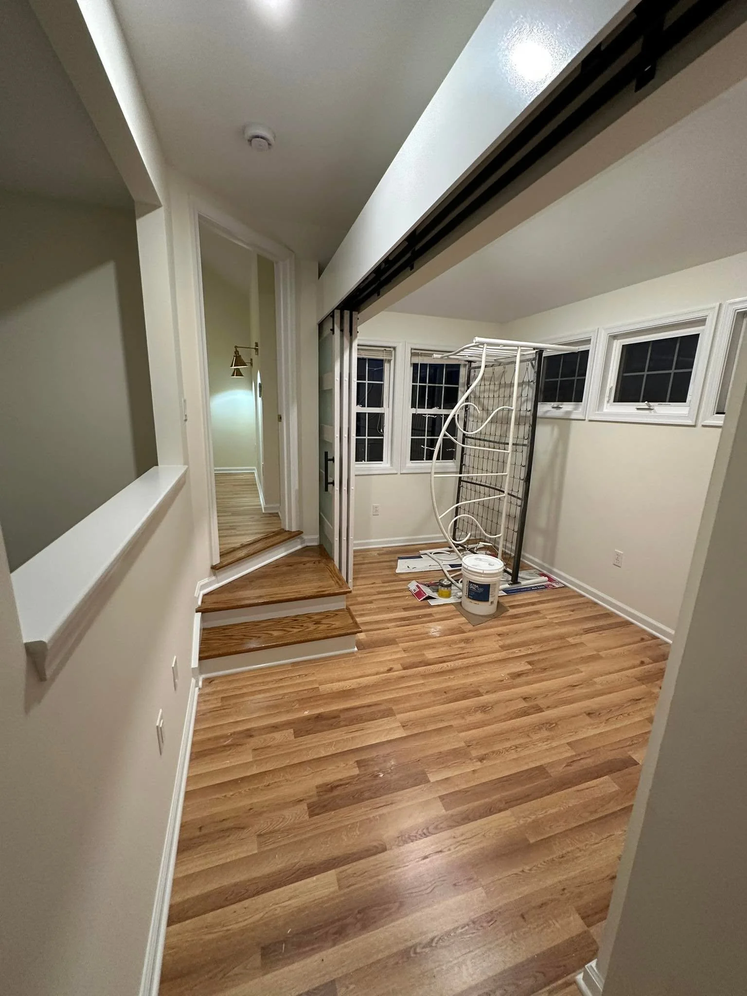 Room under renovation with hardwood floor, a metal shelving unit, paint bucket, and construction tools, leading to a hallway with three windows and a sitting area.