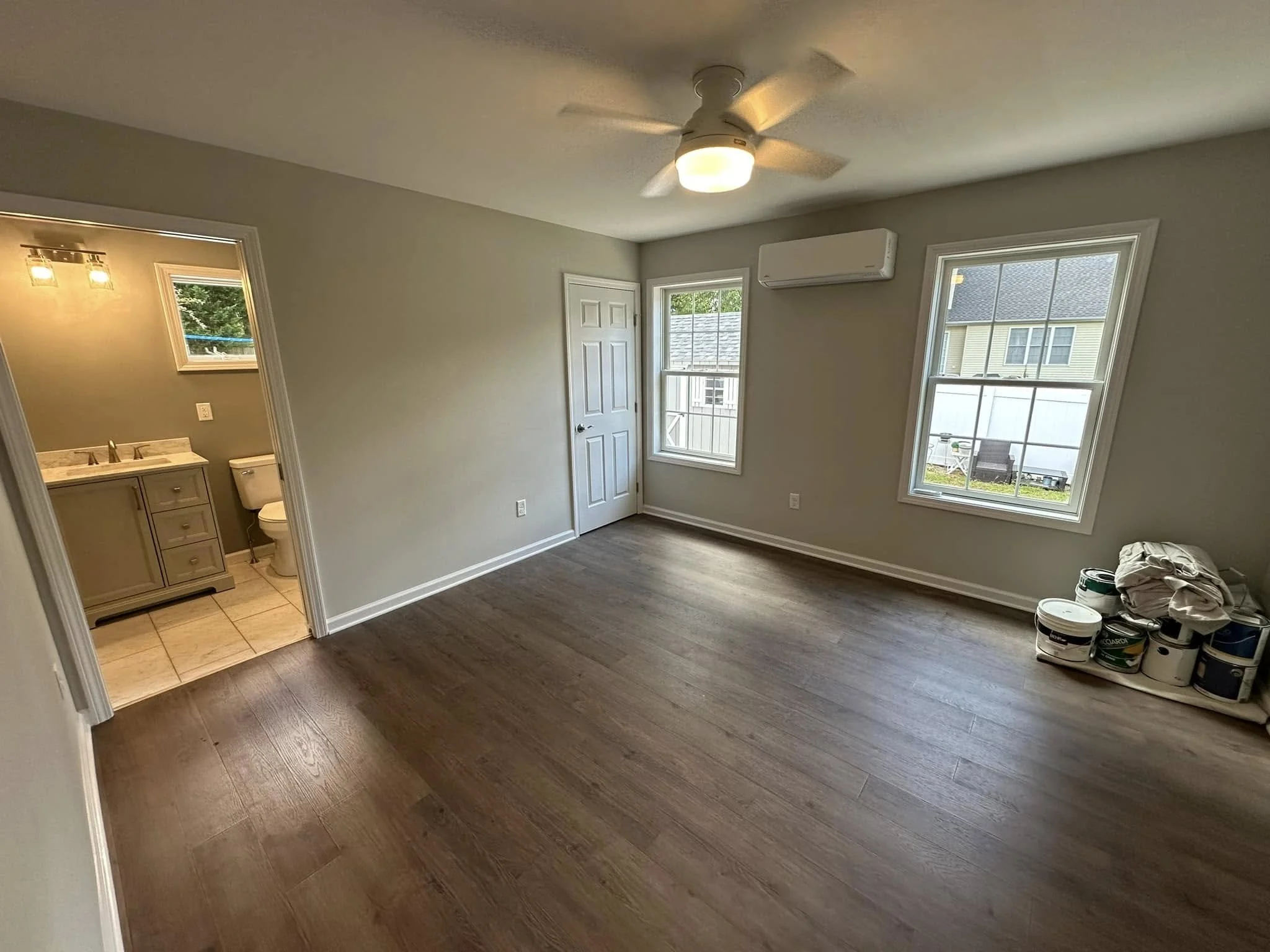 Empty room with hardwood floors, two large windows, a white door, and a ceiling fan with light. Paint cans and construction materials are on the floor, indicating ongoing or recent renovation.