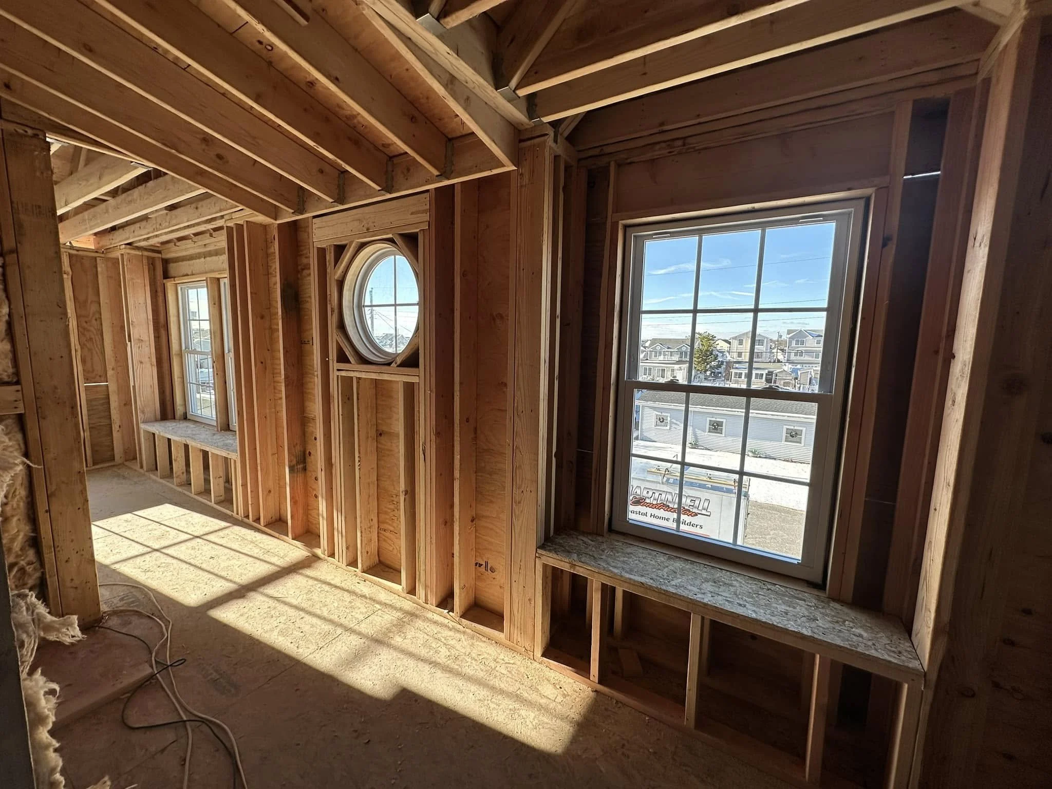 Interior of a house under construction showing wooden framing with large windows revealing a neighborhood outside on a sunny day.