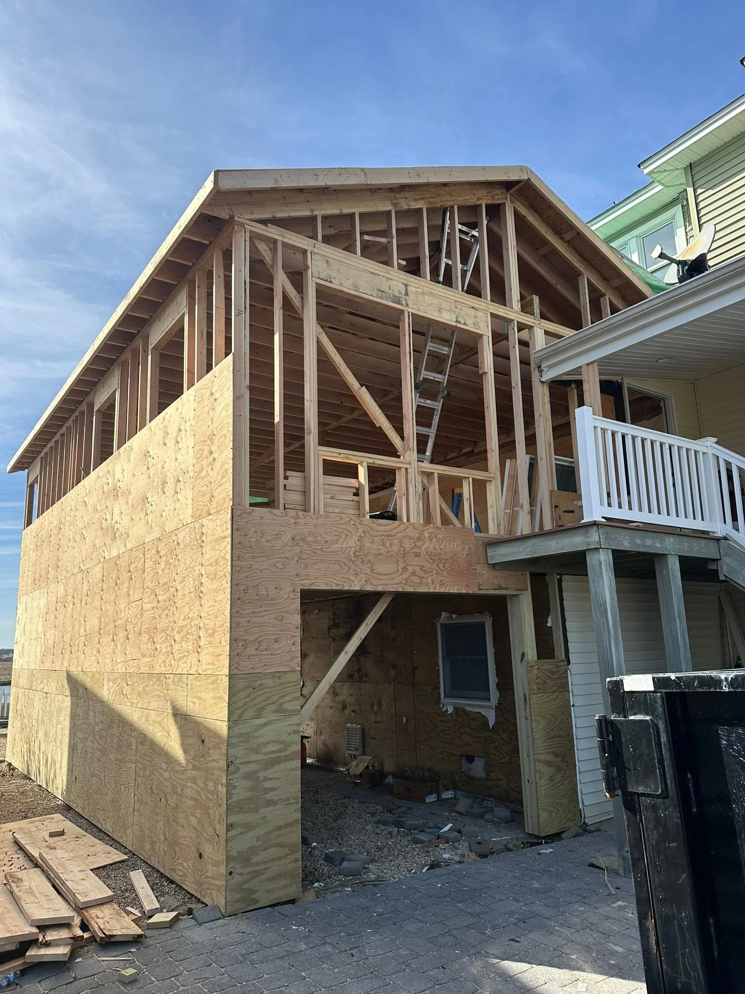A house under construction with exposed wooden framing and plywood sheathing, with a ladder leaning against the structure and construction materials scattered around.