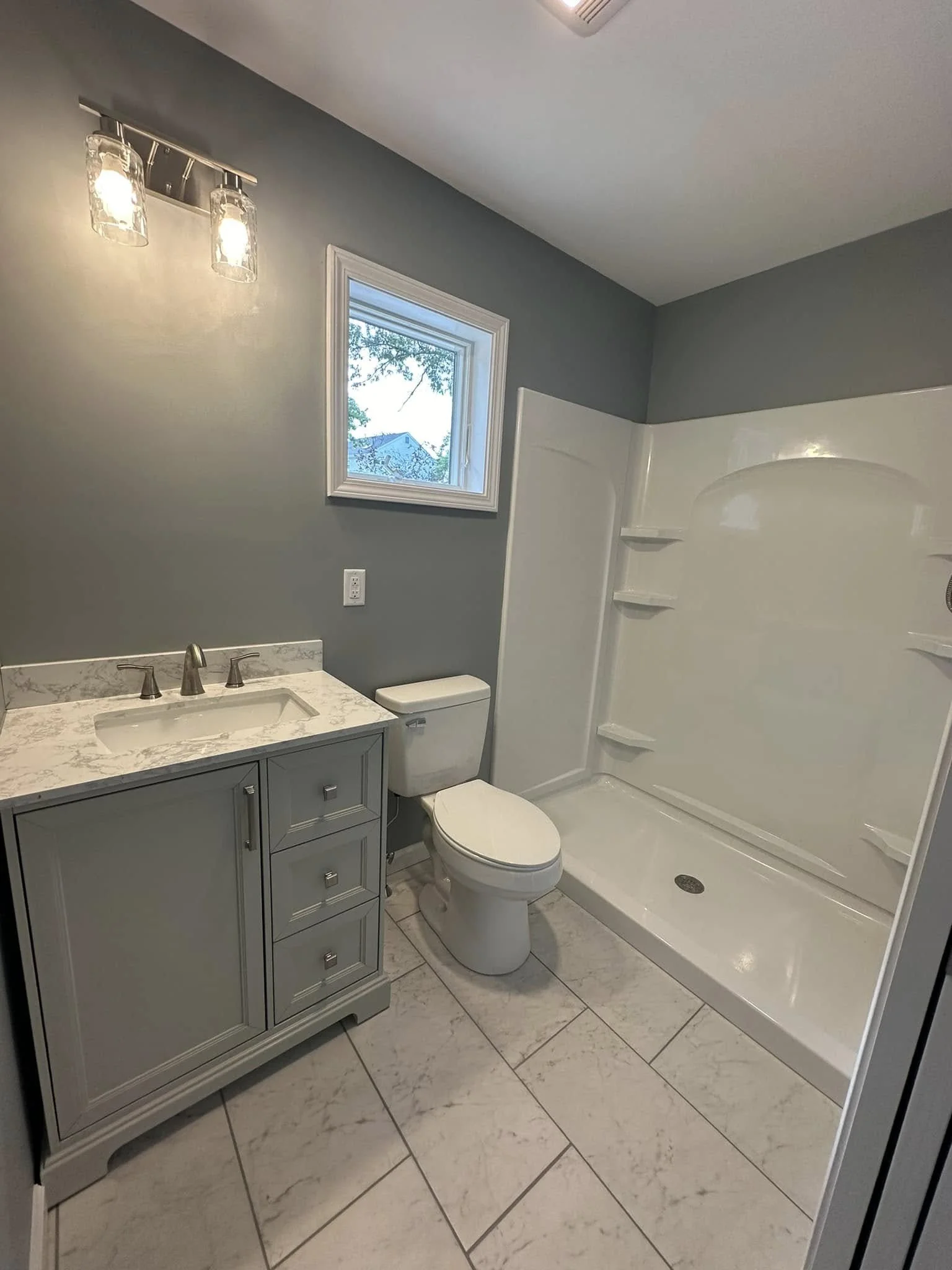 A bathroom with a gray vanity, white marble countertop, sink, metal faucet, toilet, white shower enclosure with shelves, a small window, and beige tile flooring.