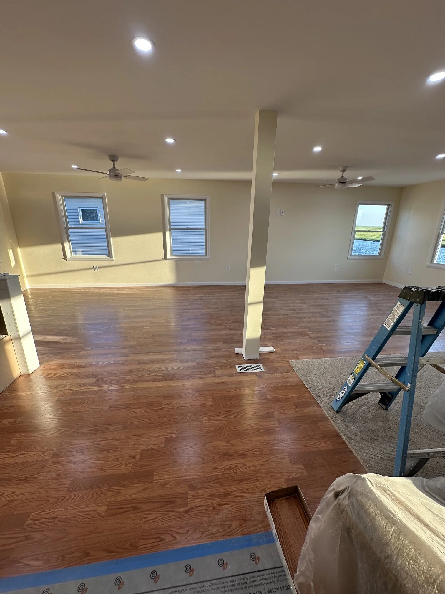 Living room with wood flooring, two ceiling fans, and multiple windows showing outdoor water and landscape. A ladder and some items are in the room, indicating ongoing work.