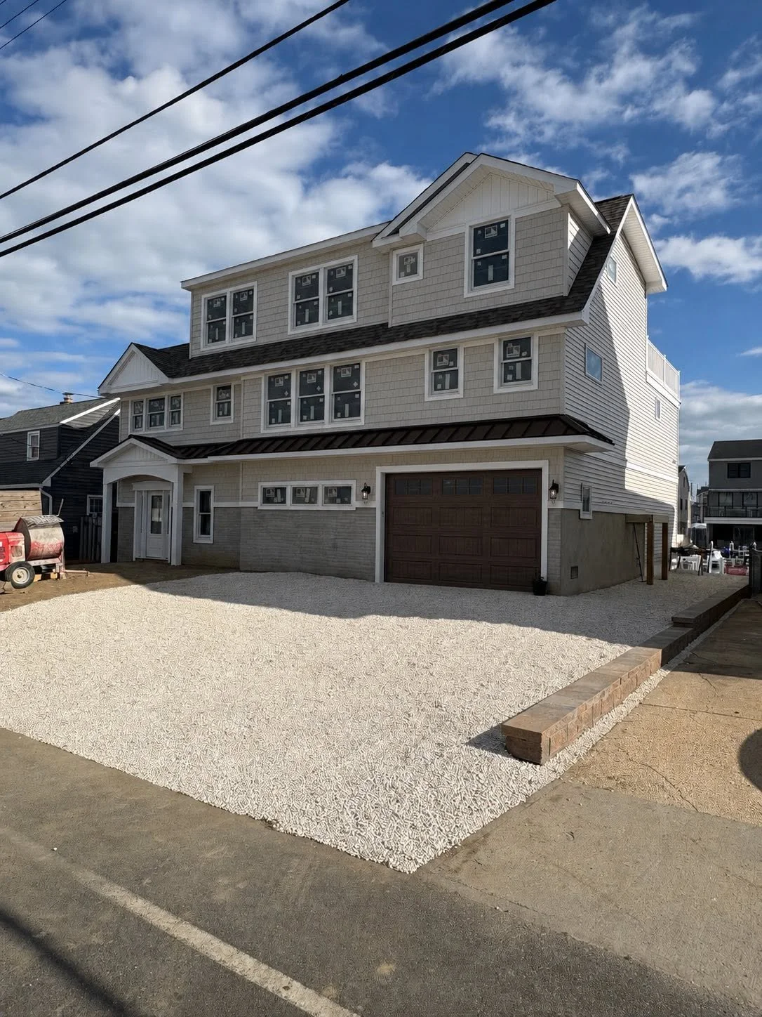 A multi-story house with light-colored siding and a dark garage door, set against a blue sky with clouds, with a gravel driveway and a small brick border in the front.