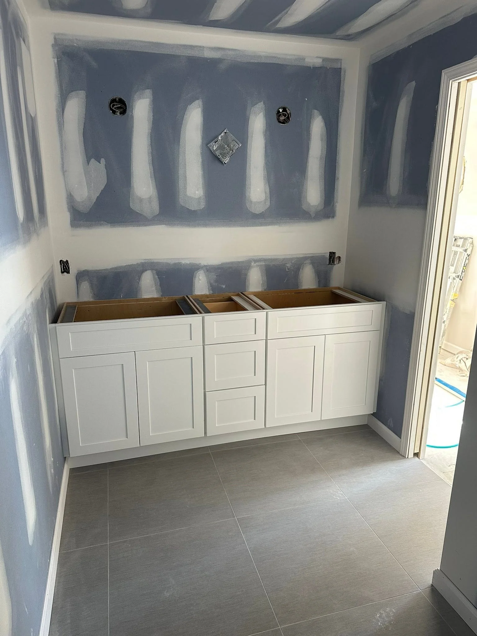 Bathroom vanity with white cabinet and drawers, gray tiled floor, blue walls, and ongoing wall painting with visible drywall patches and electrical outlets.