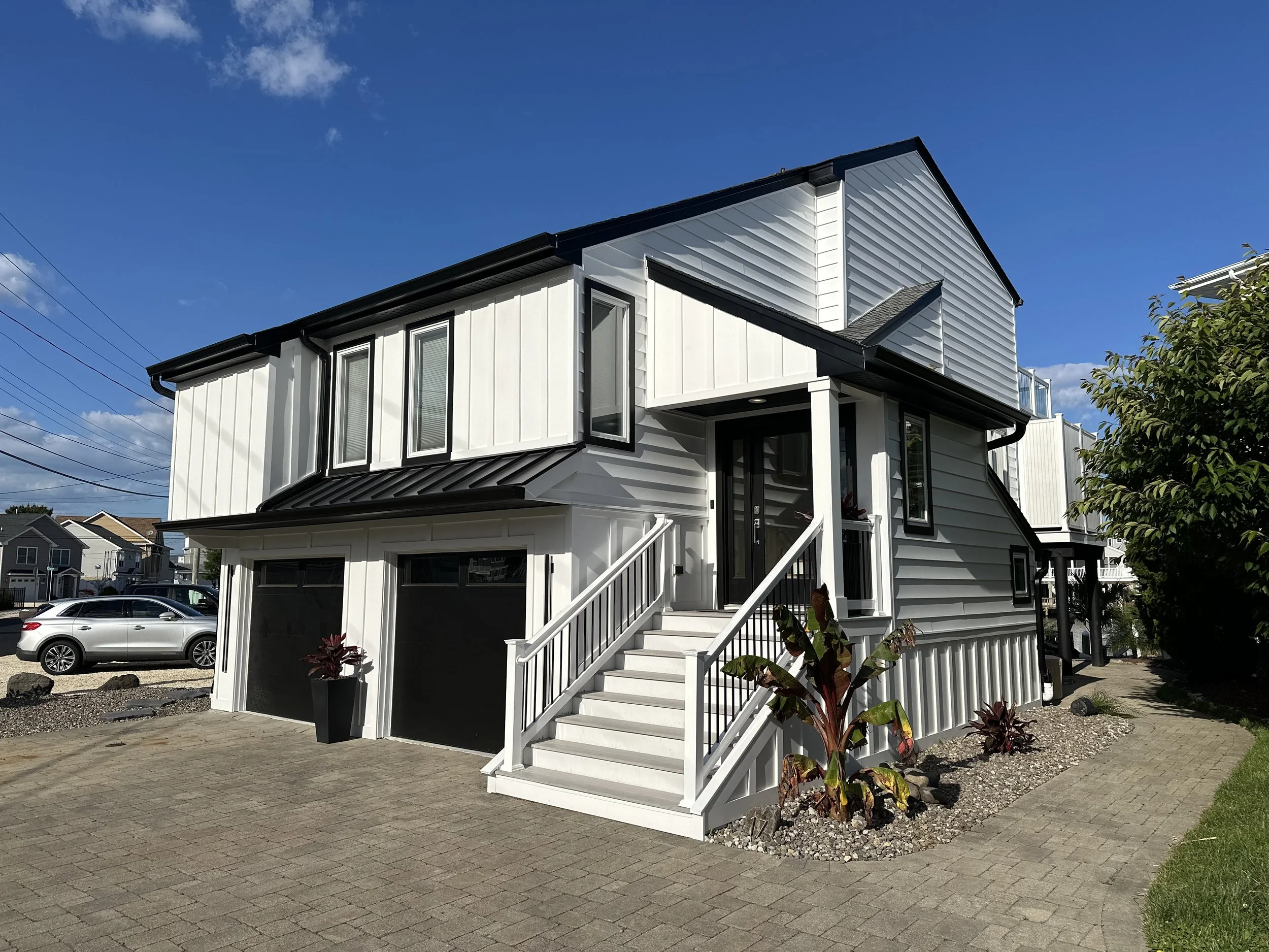 Modern white house with black trim, two-car garage, front staircase, and landscaped yard under a blue sky.