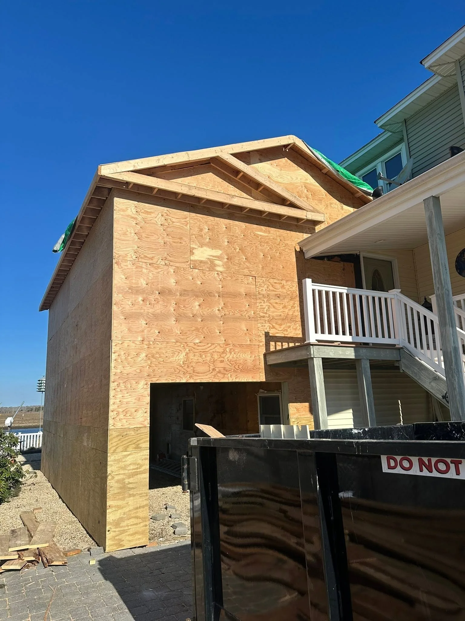 Construction site of a new building with wooden framing and plywood walls, partially finished, next to an existing house with siding and a porch.