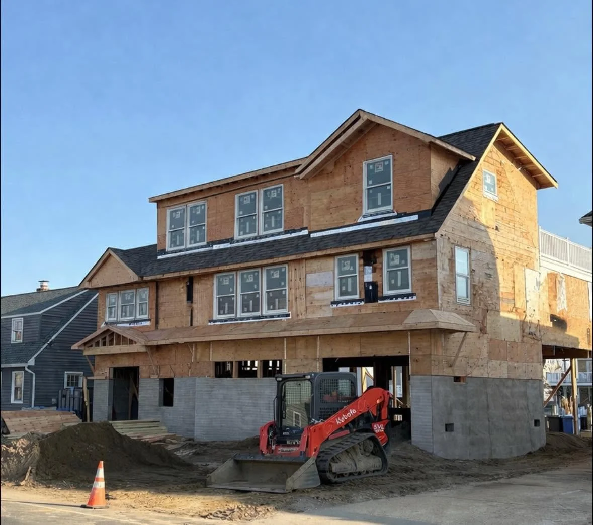 A house under construction with wooden framing and an unfinished exterior. Construction equipment and a traffic cone are in the foreground.