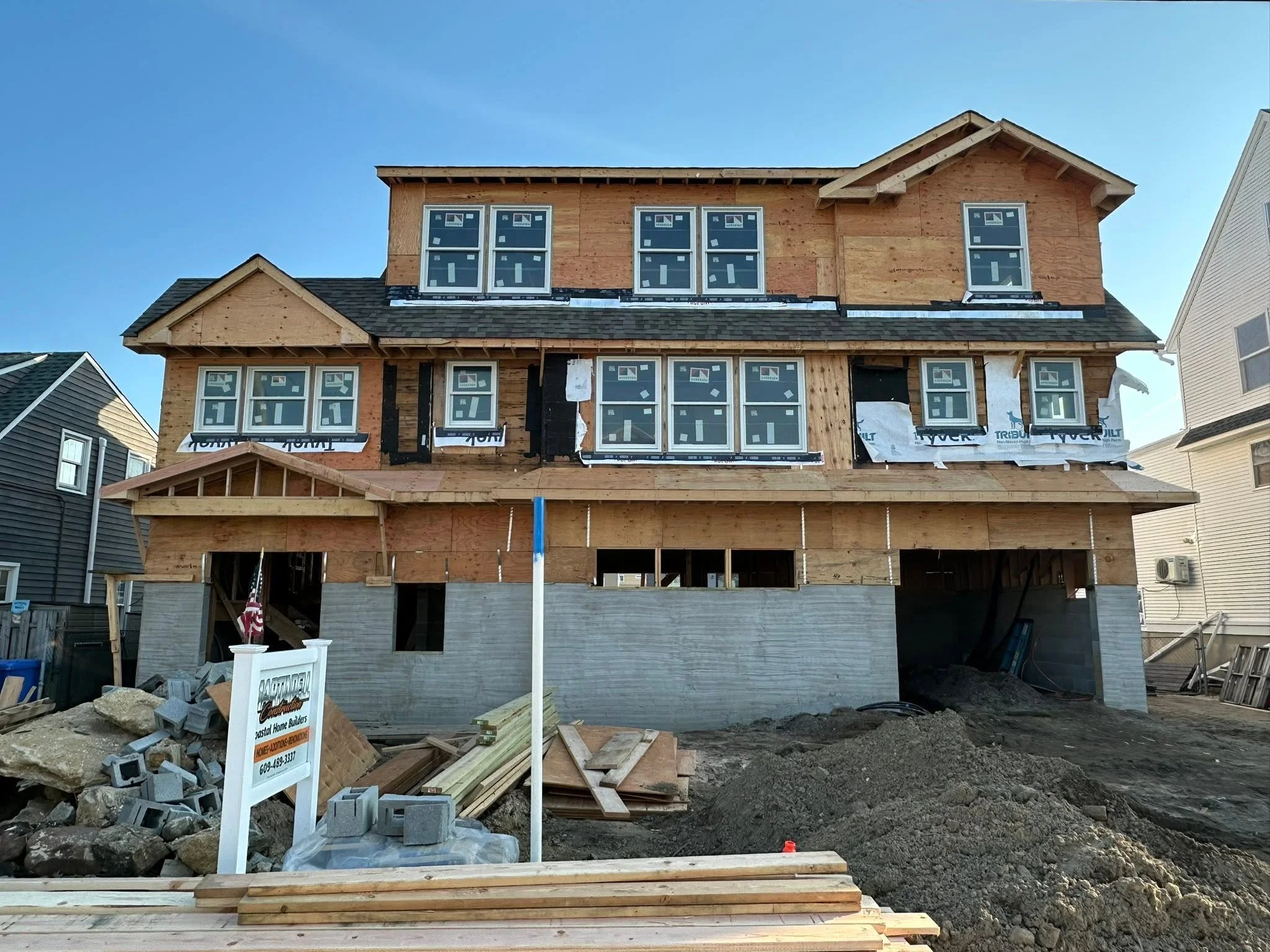 A house under construction with wooden framing, multiple windows, and a partially completed roof, surrounded by construction materials and debris.