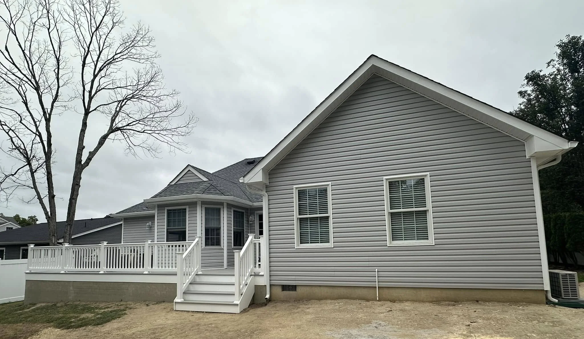 Backyard view of a two-story house with light gray siding, white trim, multiple windows, and a raised deck with white railing, with a large leafless tree and neighboring houses visible in the background.