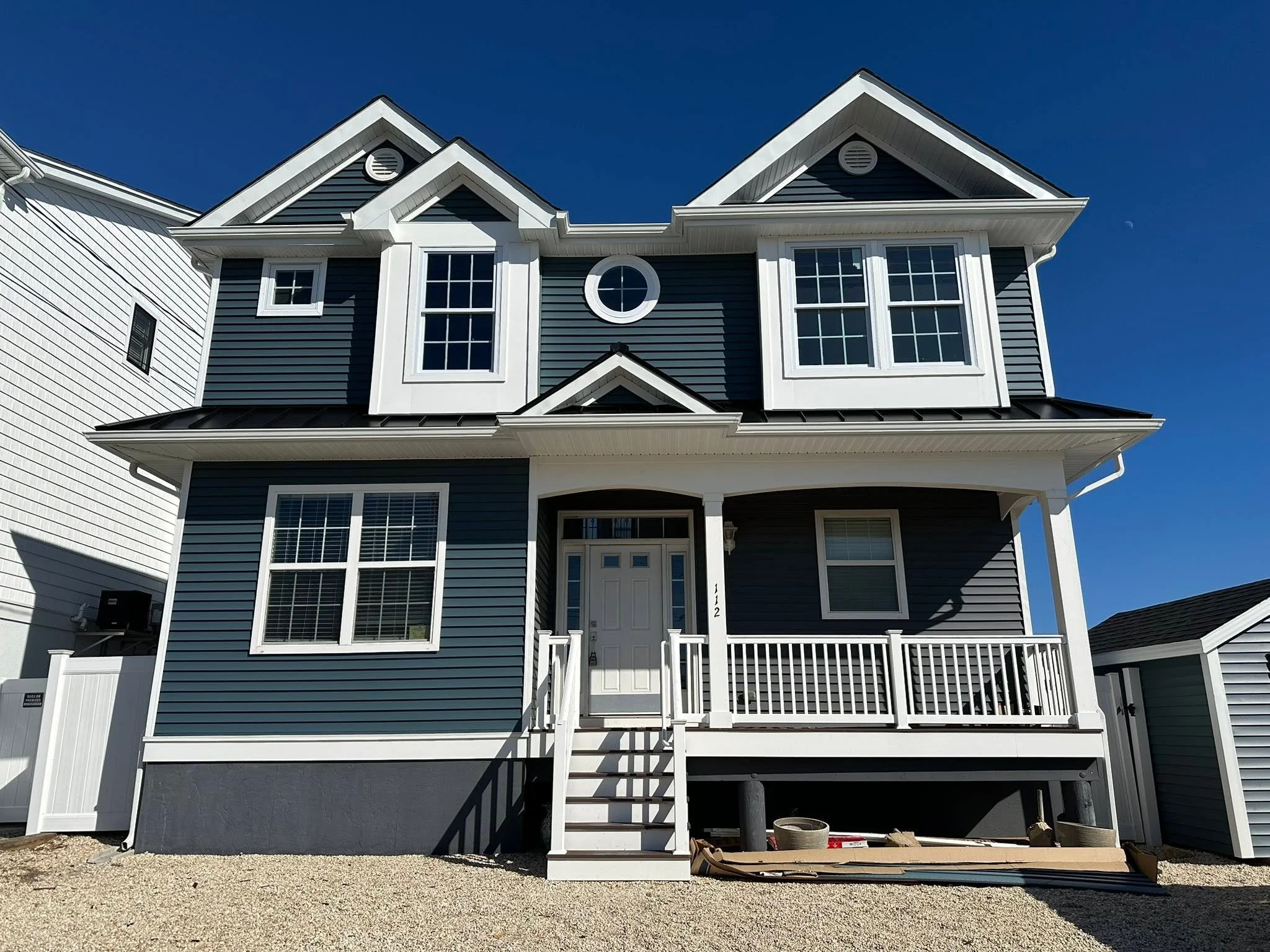 Front view of a modern two-story house with dark blue siding, white trim, large windows, a small porch, and a staircase leading to the front door, under a clear blue sky.