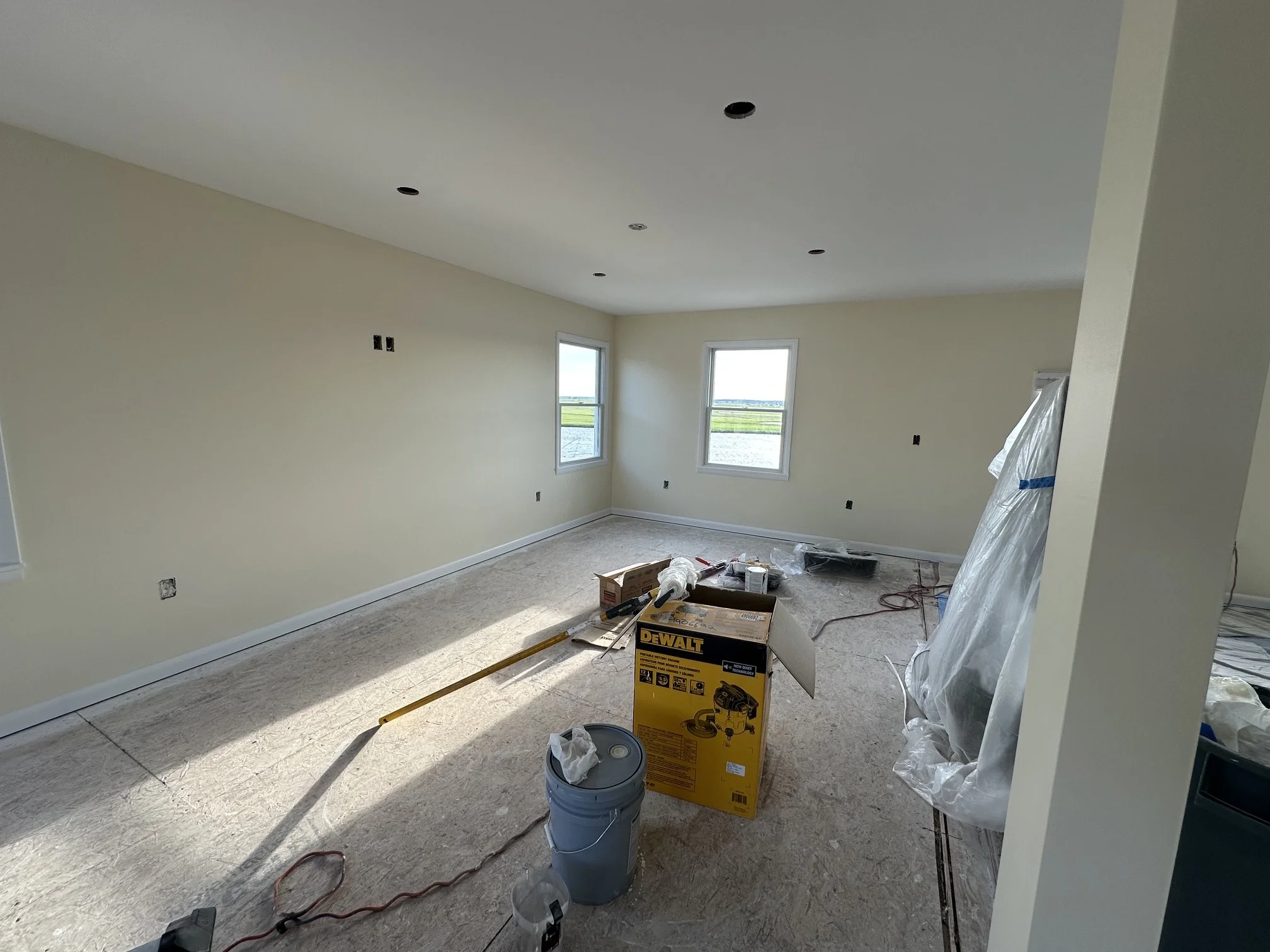 Living room under renovation with drywall installed, electrical outlets and switches not yet installed, windows letting in natural light, construction tools such as a box, bucket, and painter's drop cloth on the floor.