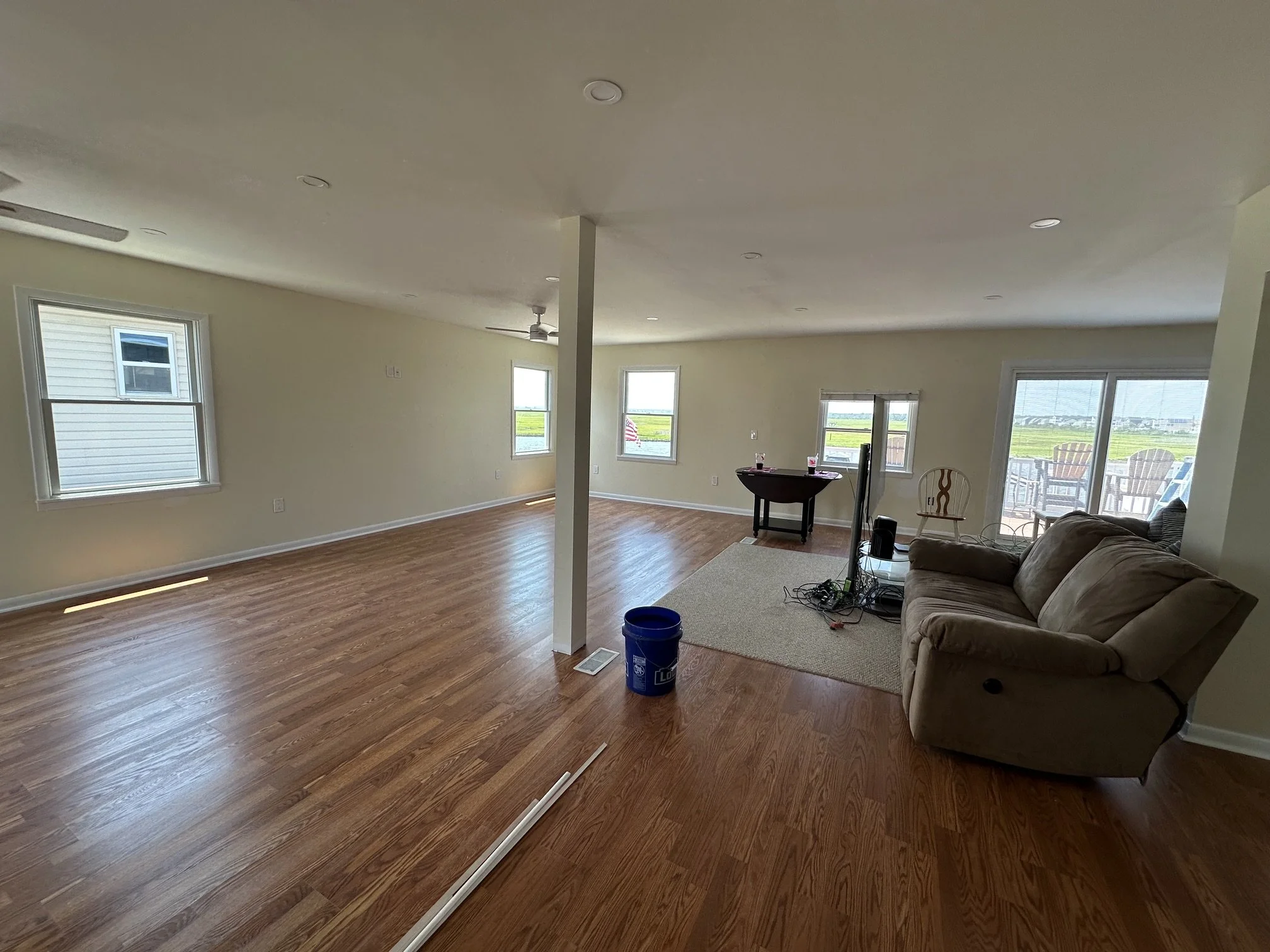 Interior of a living room with hardwood floors, beige walls, multiple windows, a ceiling fan, a brown couch, and sliding glass doors leading outside. There is a small table and some electronic equipment on the carpeted area.
