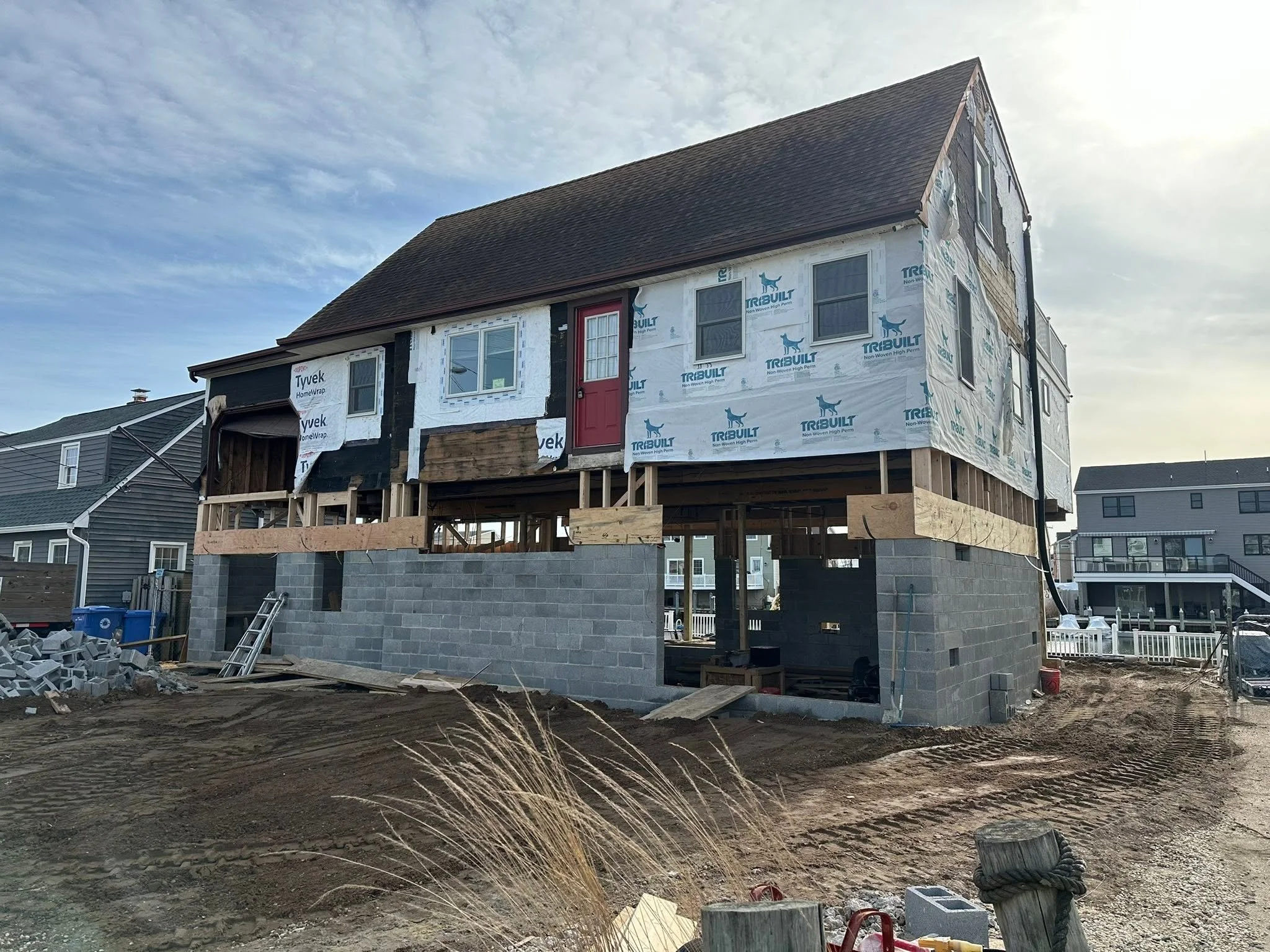 A house under construction with exterior walls covered in insulation and sheathing, elevated on concrete blocks with construction materials and tools around the site.