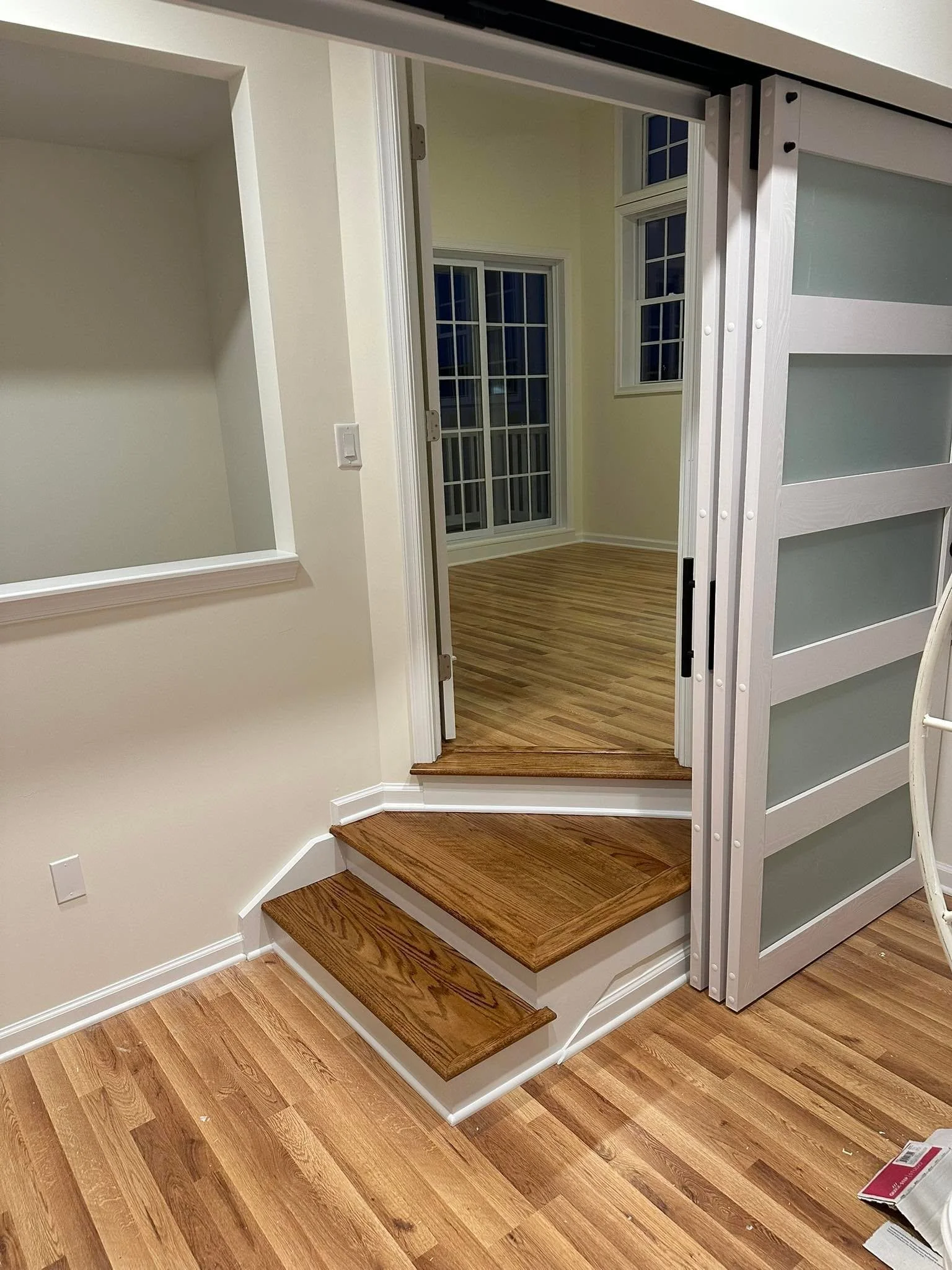 Interior of a house showing a wooden staircase with three steps, hardwood floor, an open sliding door with frosted glass panels, and a room with hardwood flooring and multiple windows.