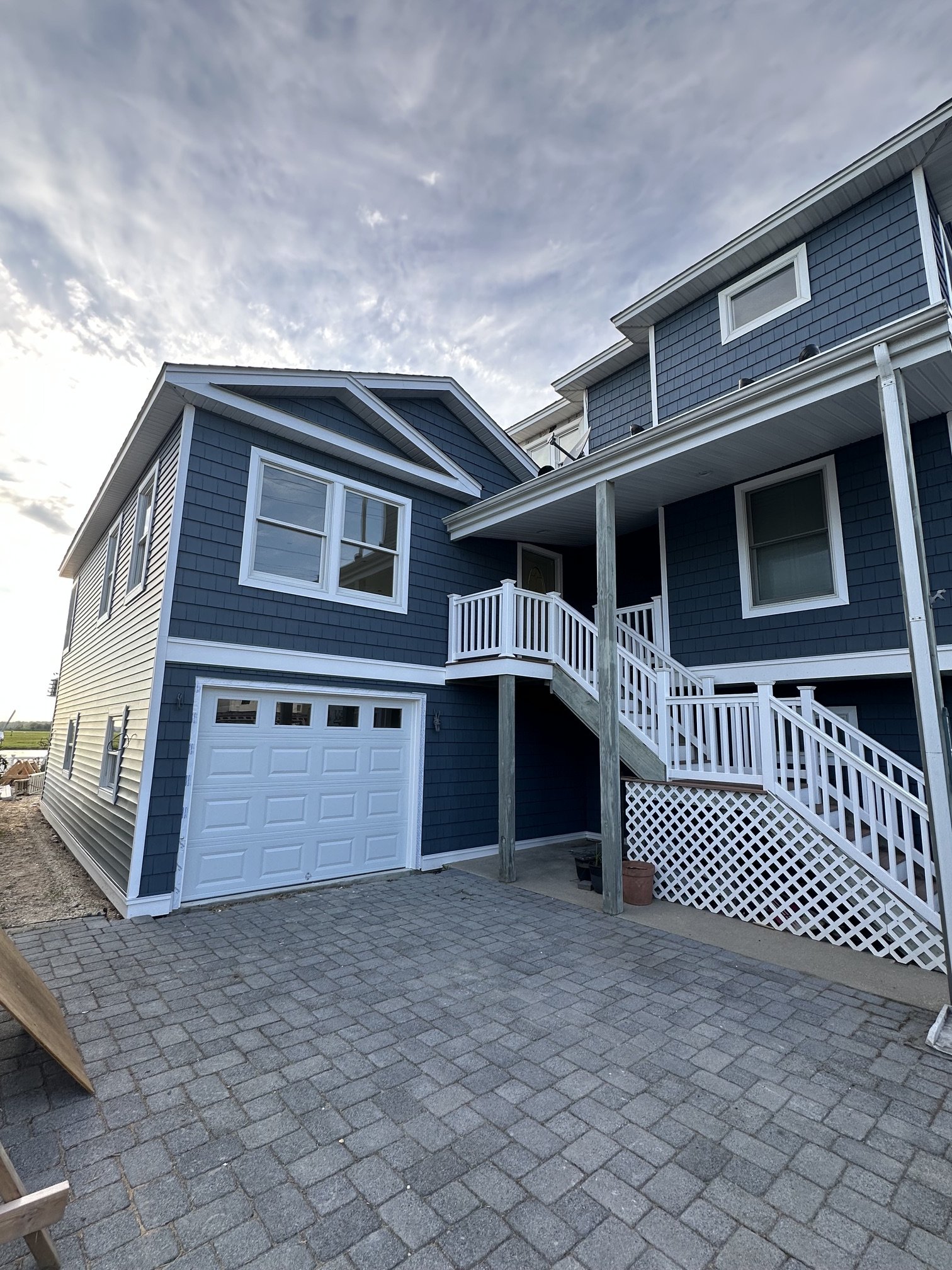 A blue two-story house with whitetrim, a garage with a white door, and a deck with stairs leading up, with a brick patio in the foreground under a partly cloudy sky.