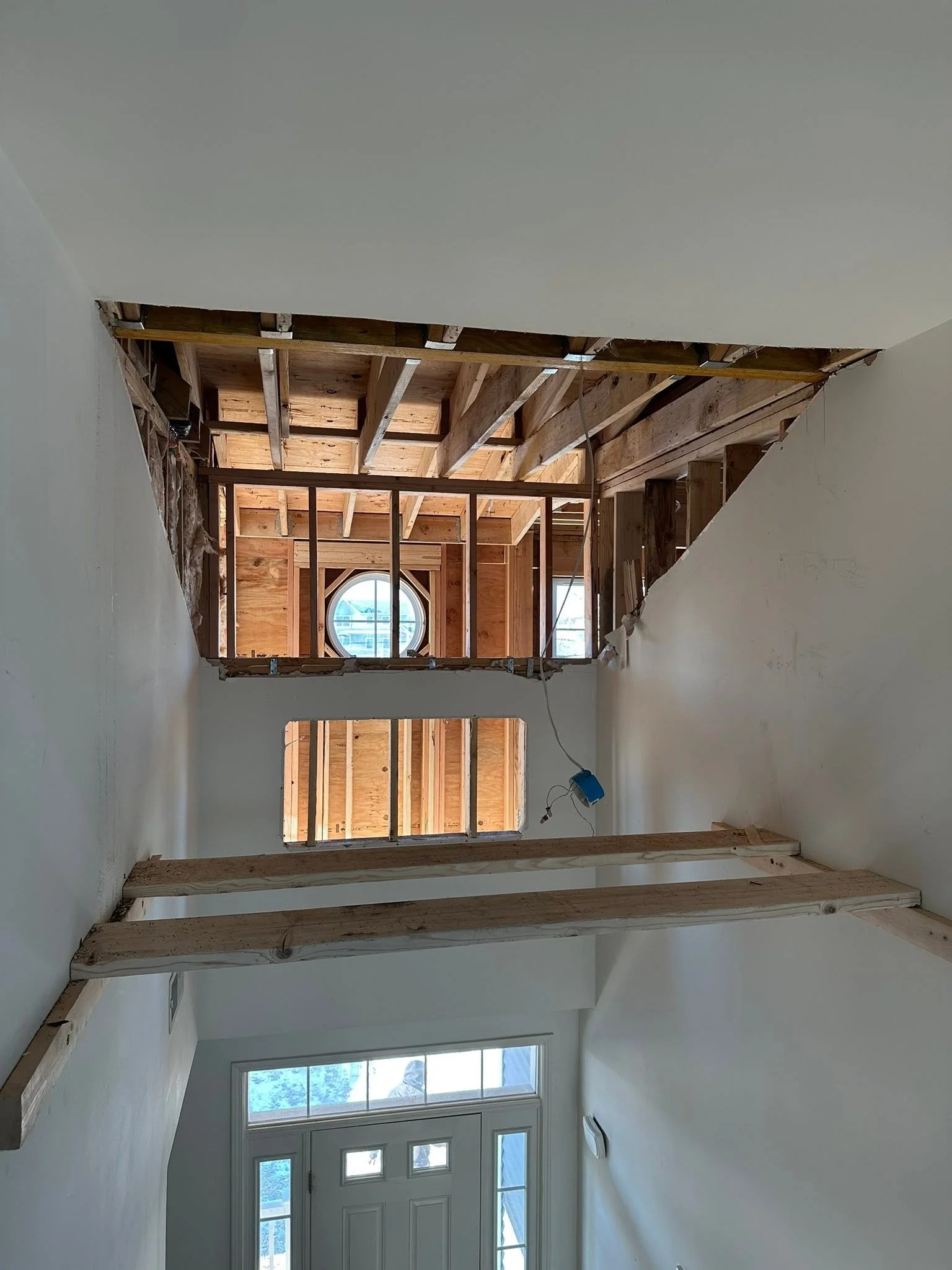 Interior view of a room under construction, showing exposed wooden framework for walls and ceiling, with a building visible through windows at the front.