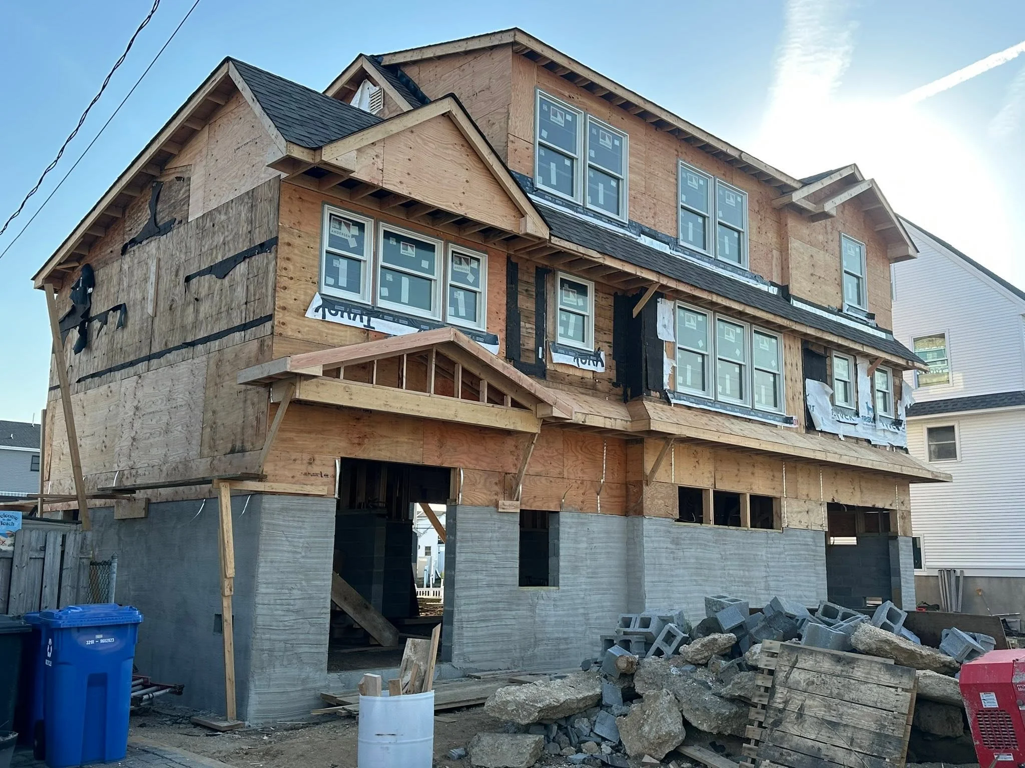 A multi-story house under construction with wooden framing and windows installed, surrounded by construction debris, rocks, and construction equipment.