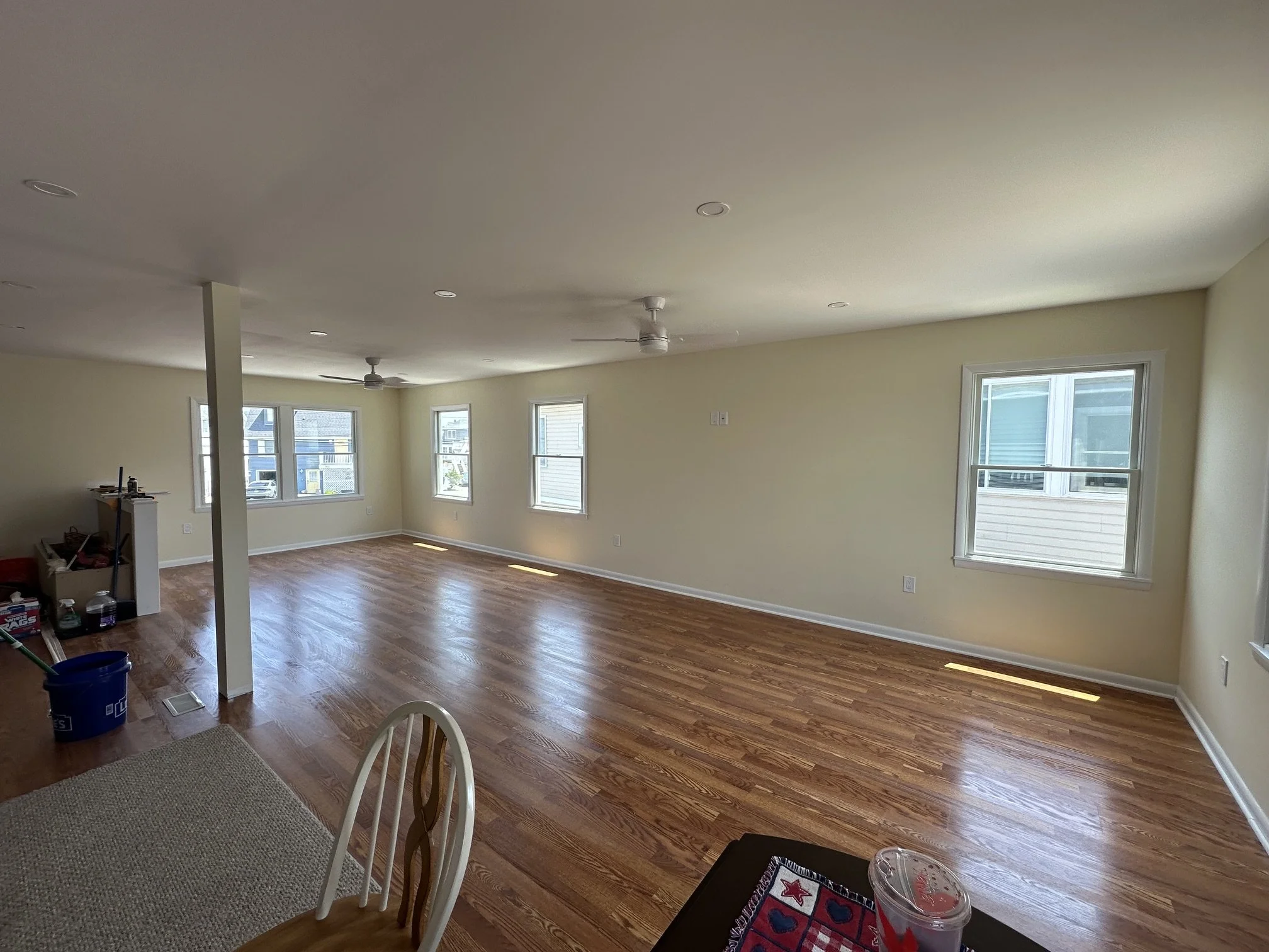 Empty living room with large windows, hardwood floors, ceiling fans, and light-colored walls. There is some clutter near a small wall, and a table with drinks and a decorative mat in the foreground.
