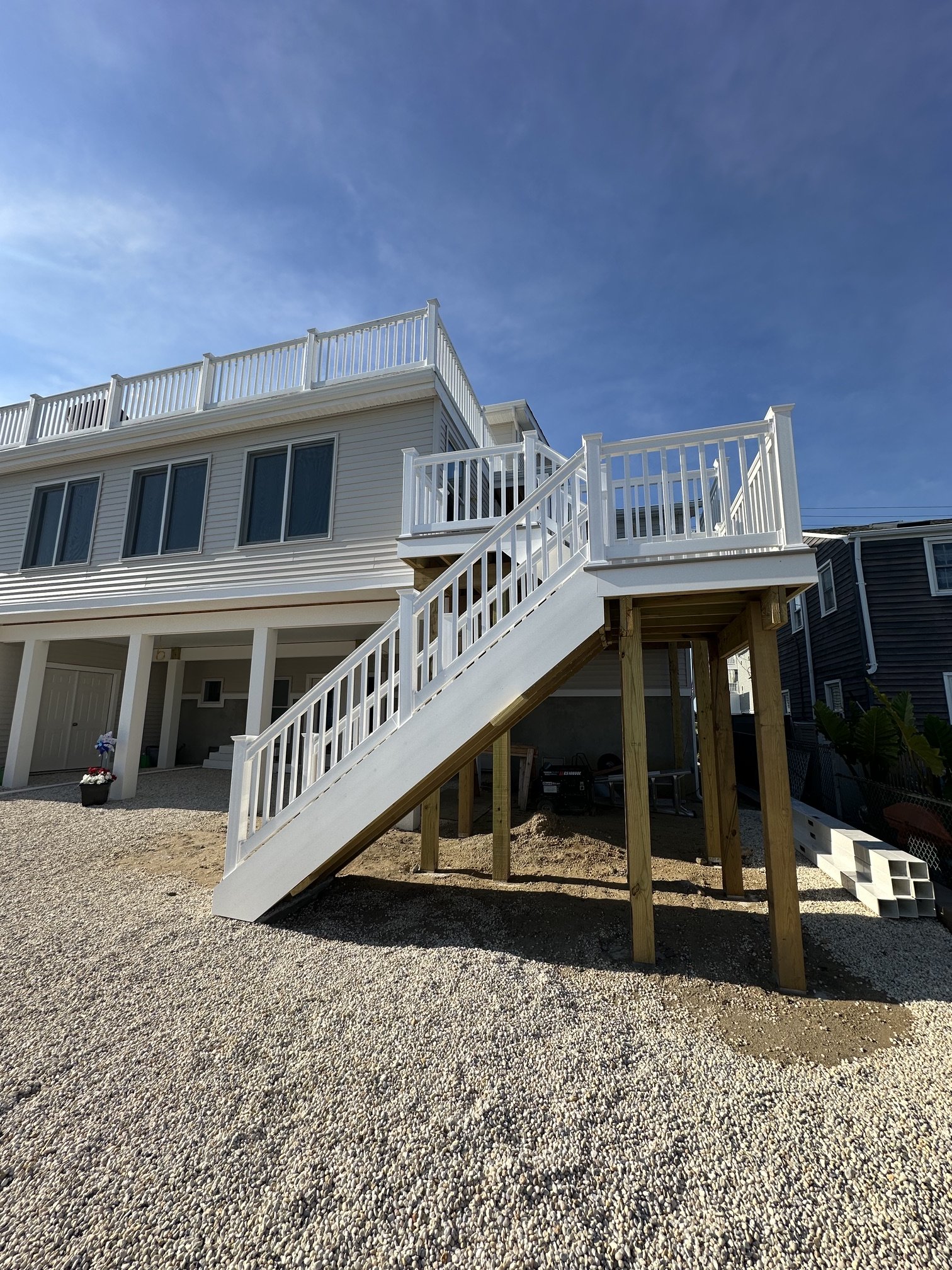 A two-story house with white siding, large windows, and an outdoor staircase leading to a second-floor balcony, with gravel ground in the foreground.