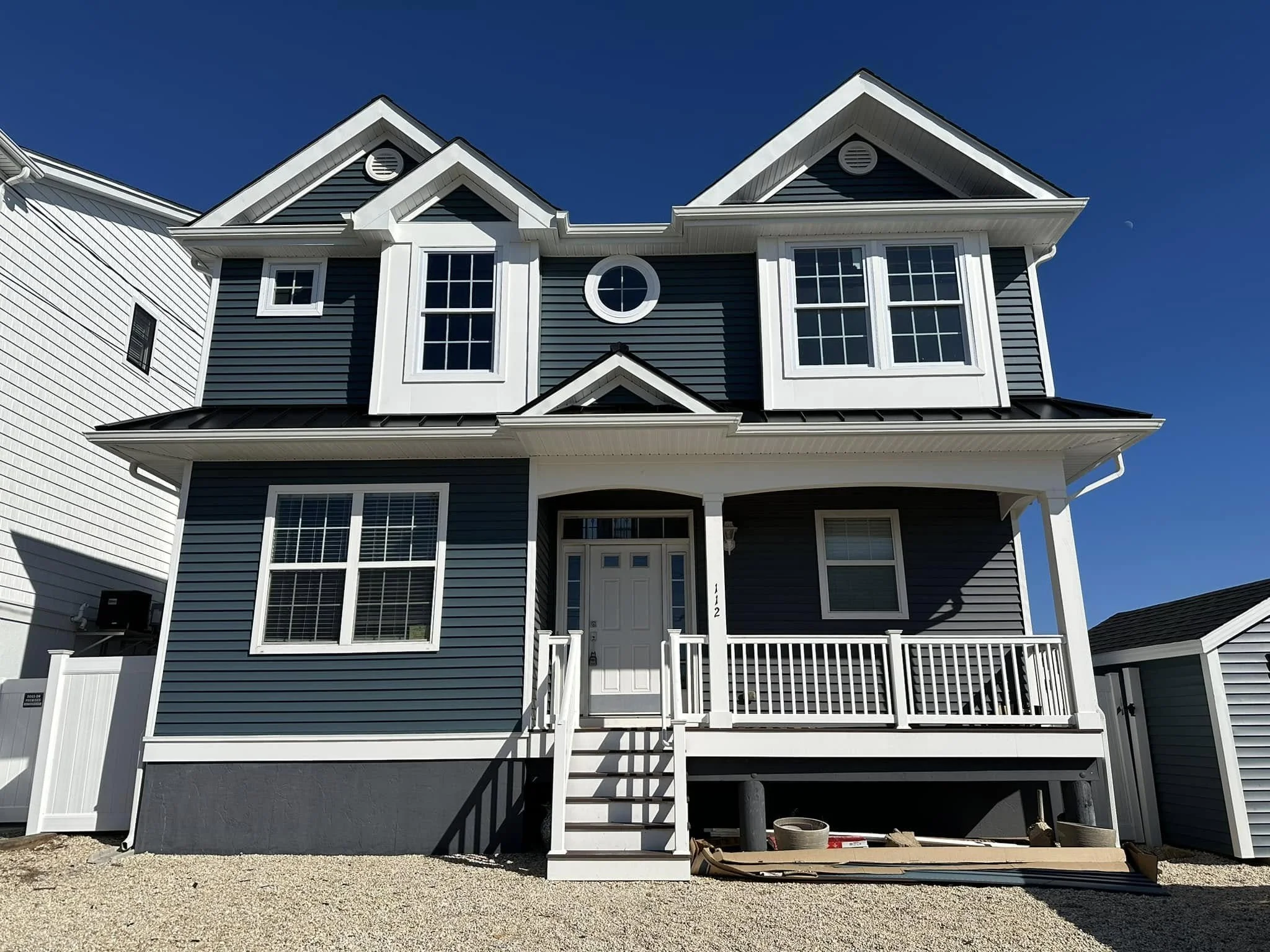 Front view of blue and white house under construction, with white railing, stairs, and a large window, against a clear blue sky.