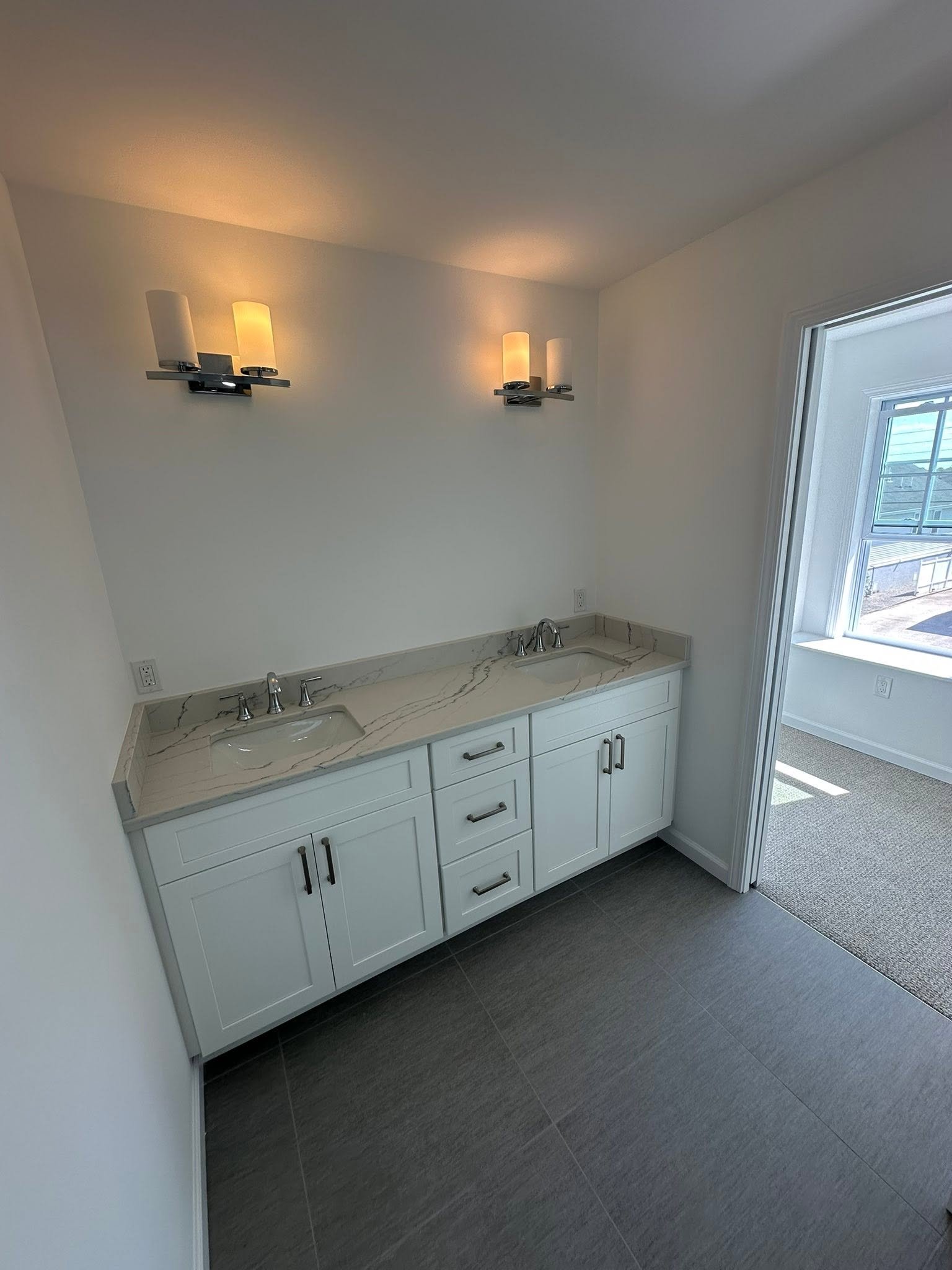 Bathroom vanity with two sinks, white cabinets, marble countertop, and a wall light fixture with four frosted glass shades illuminated. There is a window to the right showing part of the outside.