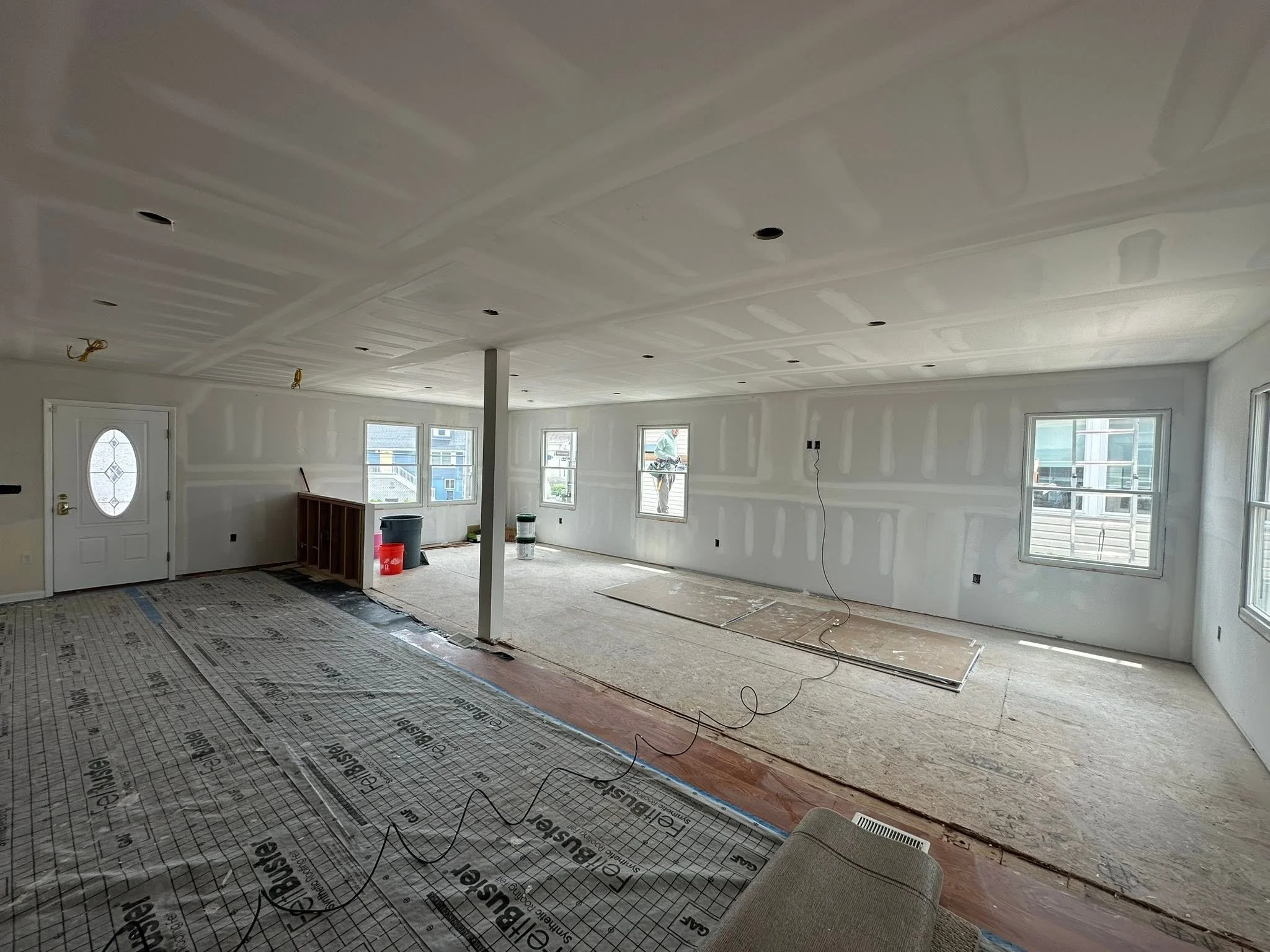 Living room under construction with drywall walls, unfinished ceiling, and flooring in progress. Construction materials and a worker visible in the background.