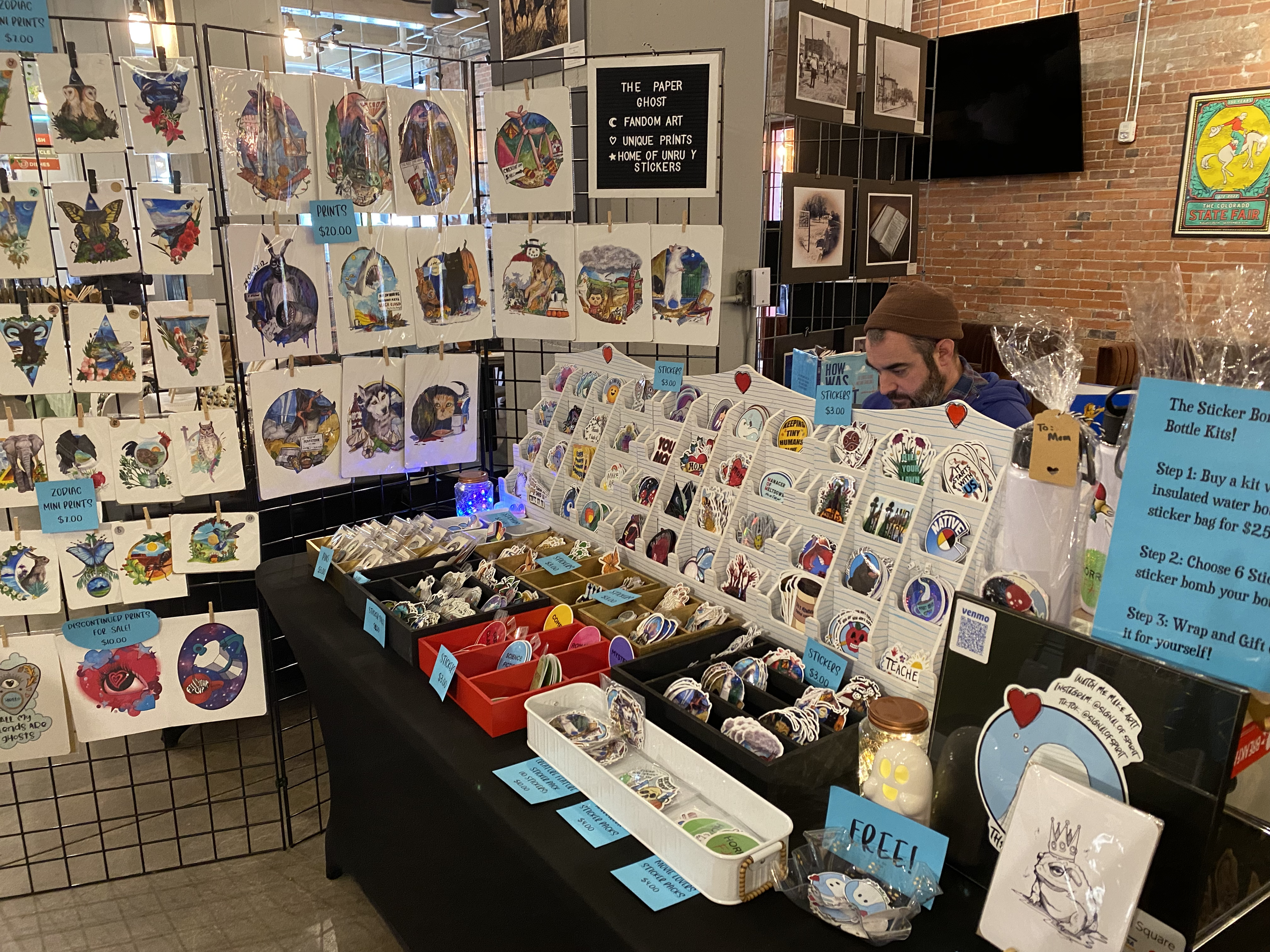 Art and sticker booth at a craft fair displaying prints and stickers. Various artwork features animals and nature themes. Pricing signs offer options for prints and sticker collection kits. A vendor is visible behind the table.