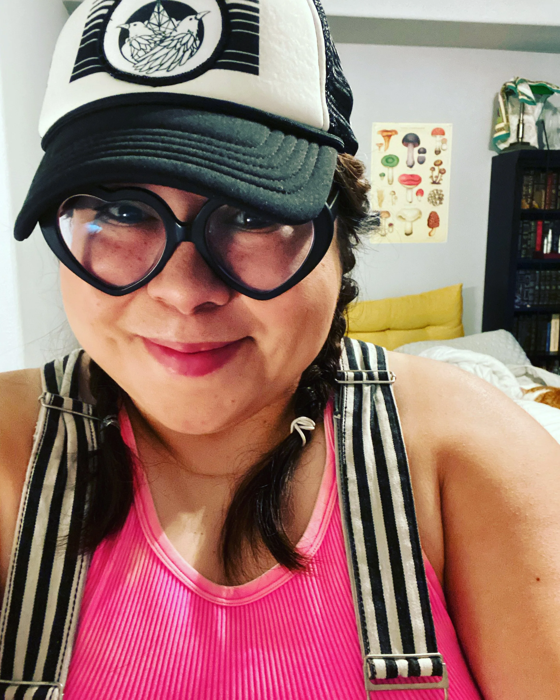 Person wearing glasses, hat, and striped overalls, with a pink top, in a room with mushroom poster and bookshelf.
