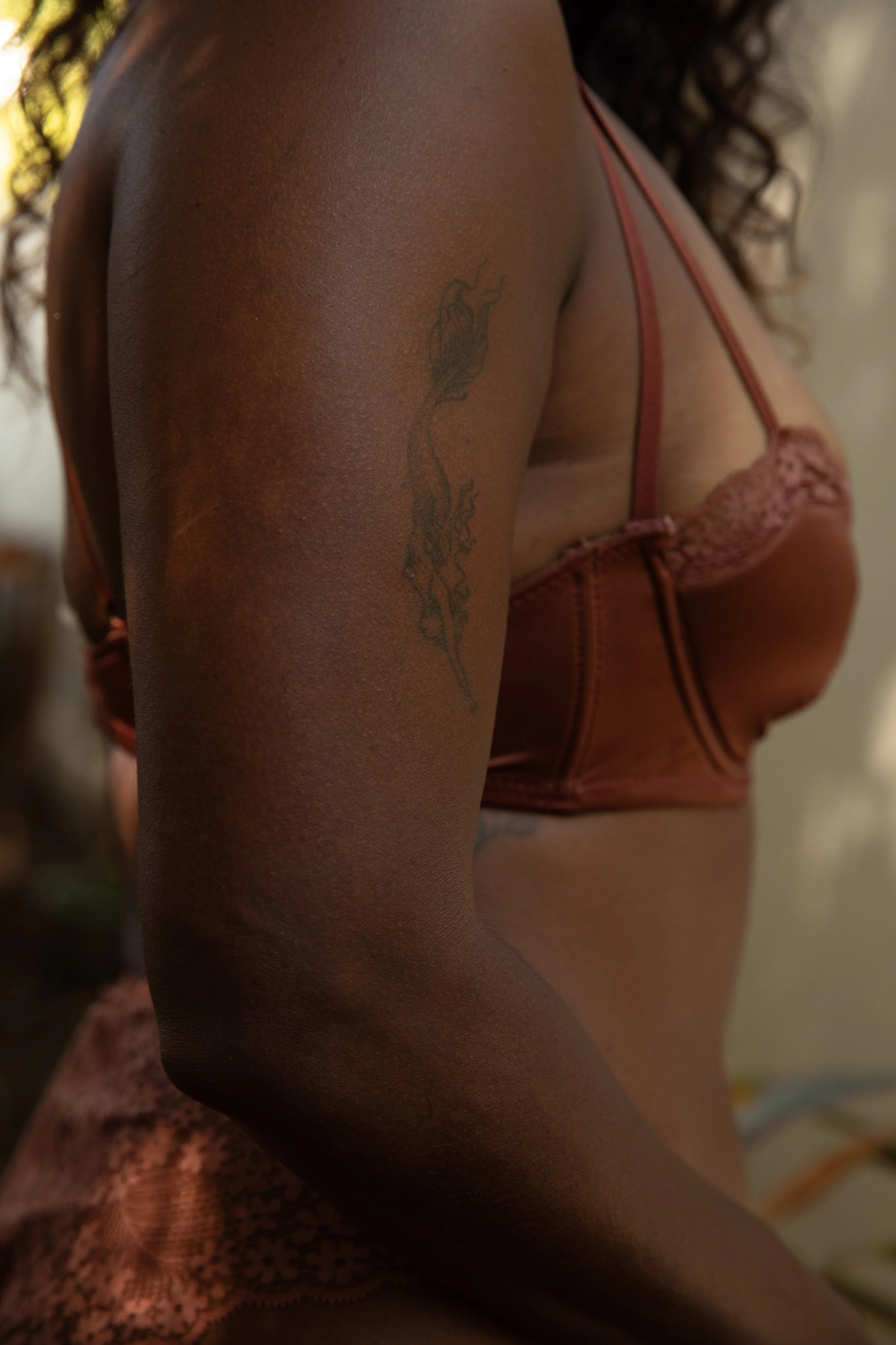 Close-up of a person's shoulder and chest area, wearing pink lace lingerie, with a tattoo on the shoulder, and curly hair visible in the background.