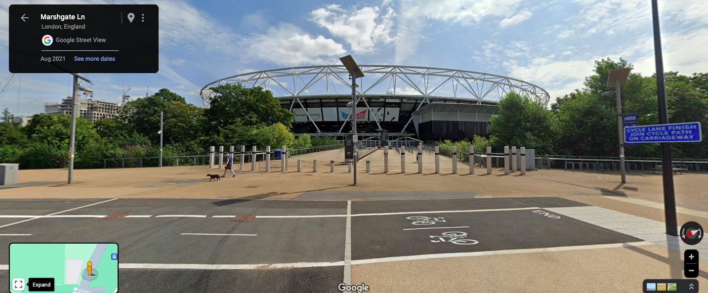 Google Street View image of a pedestrian walking dog near a stadium with a white roof structure, trees on sides, and blue sky with clouds in London's Marshgate Lane.