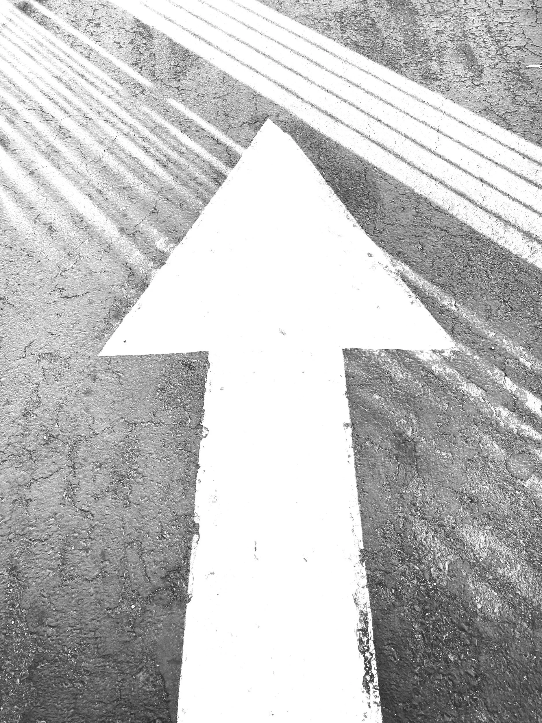 A white arrow painted on pavement, pointing straight ahead, next to textured concrete and raised tactile paving strips.